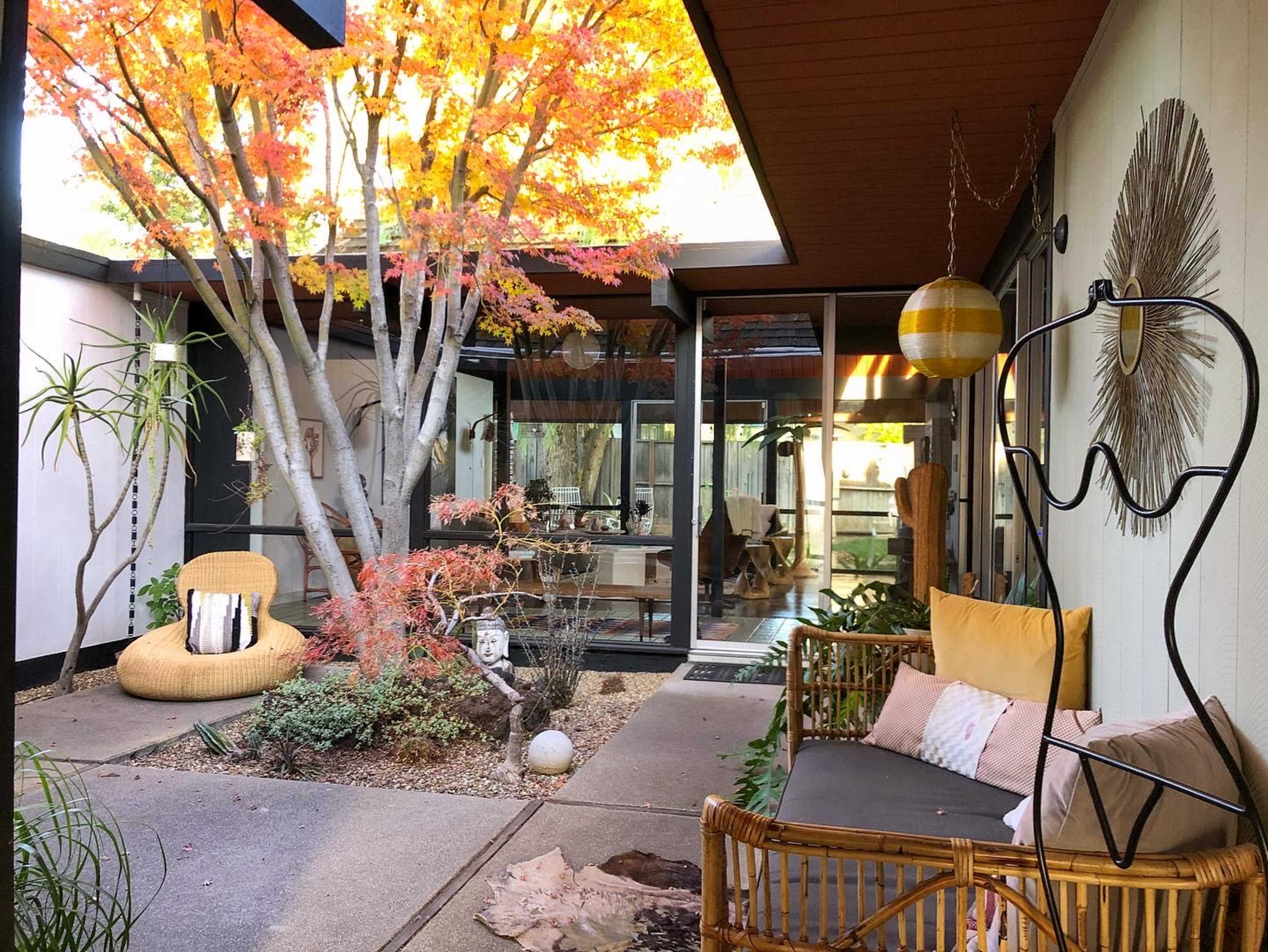 A modern courtyard with a seating area surrounded by trees and plants, featuring a glass wall that opens to an interior space.