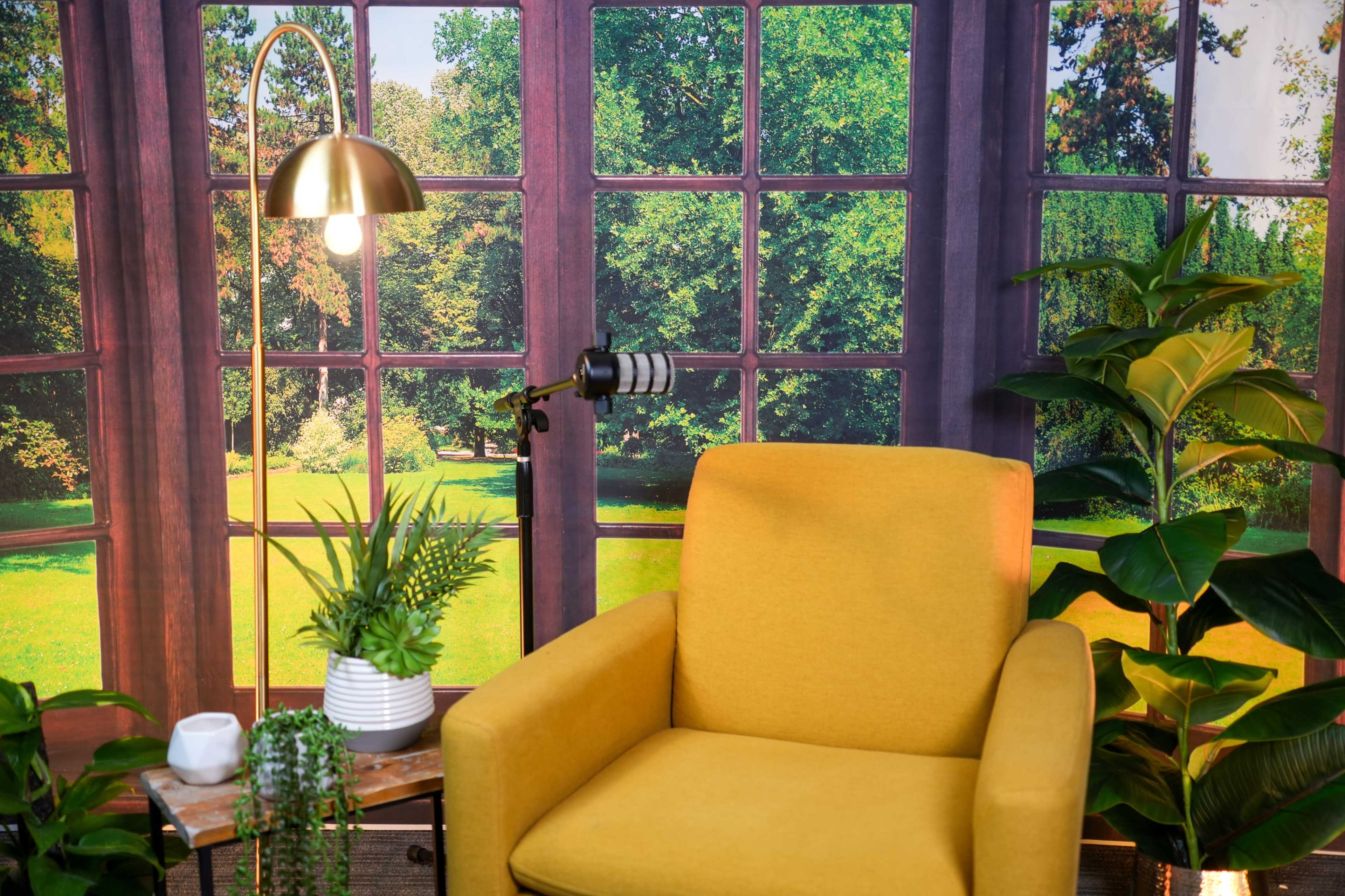 The image features a yellow armchair beside a small table with plants, set against a backdrop of large windows overlooking a green garden.