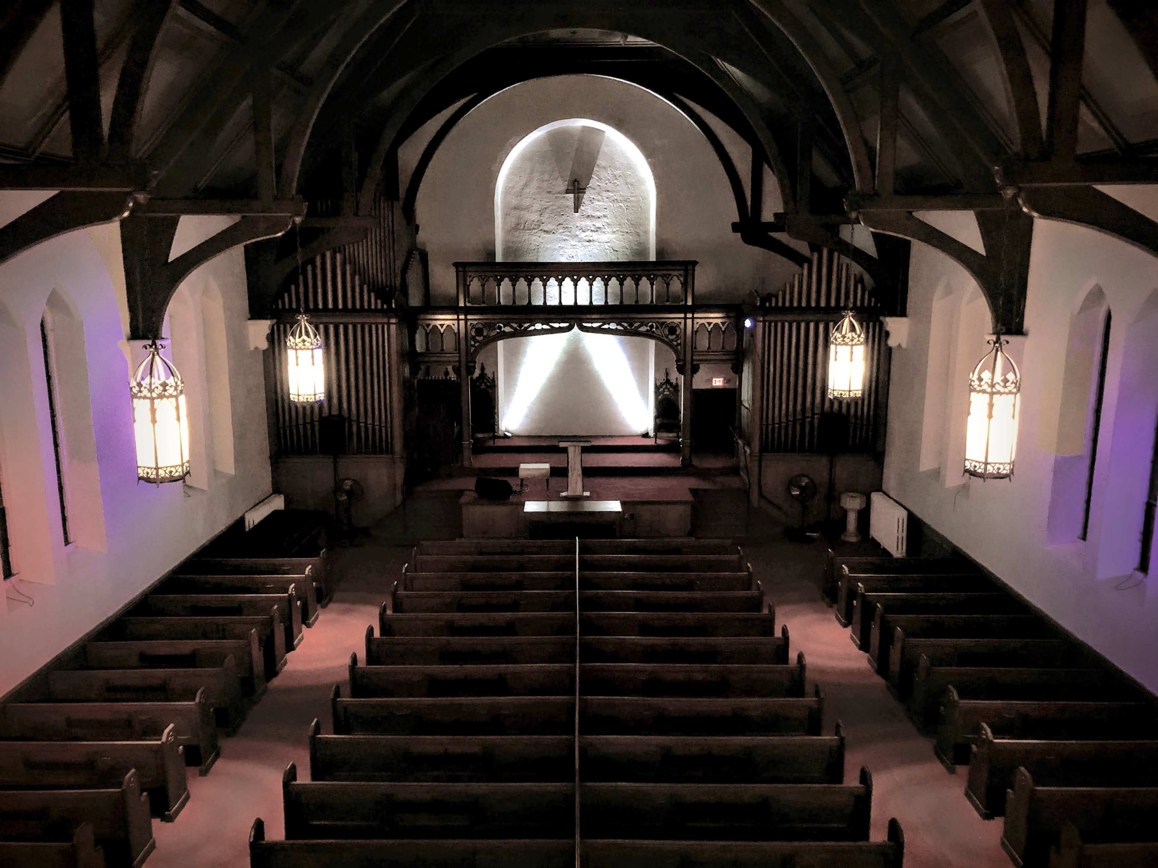 The interior of a church with wooden pews arranged in rows facing an altar at the front, illuminated by pendant lights.