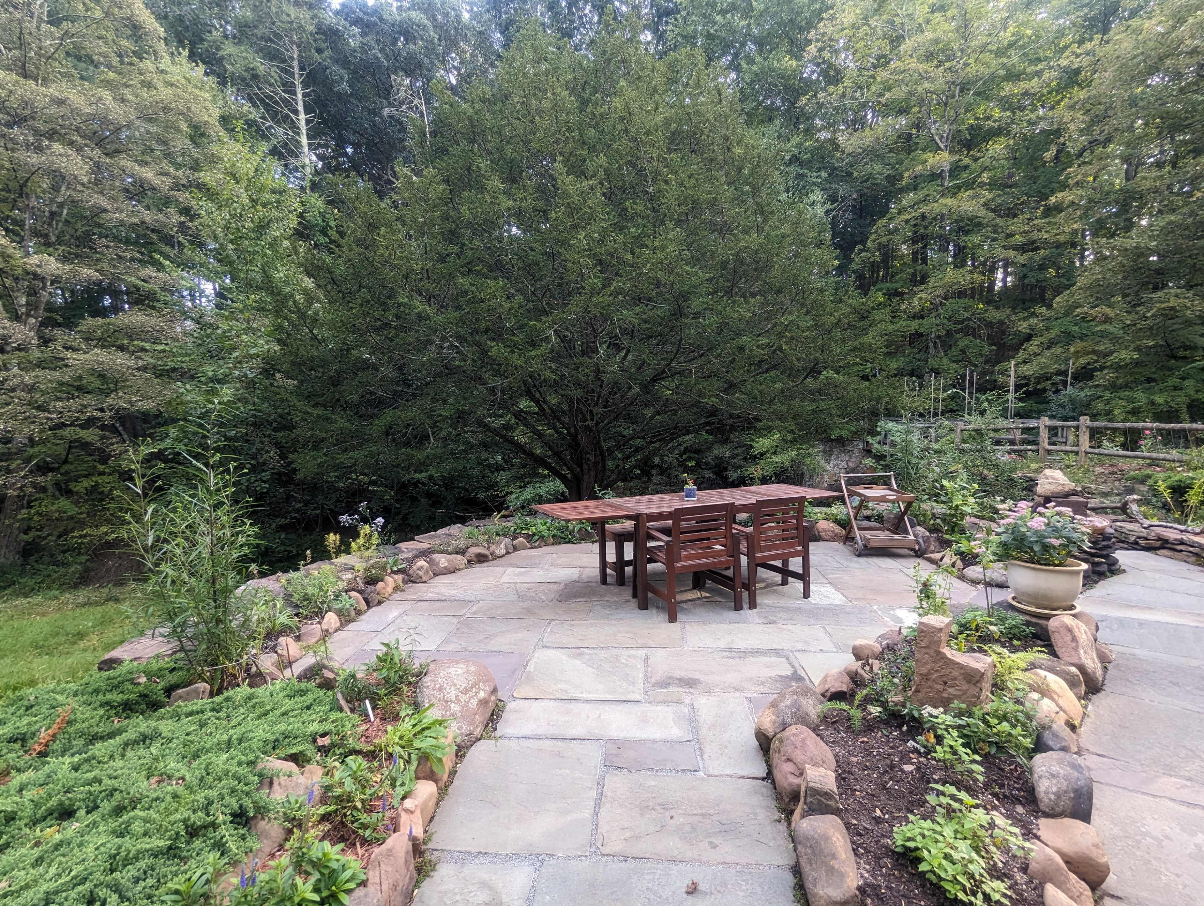 A stone patio features a wooden dining table and chairs surrounded by a landscaped garden and large trees.