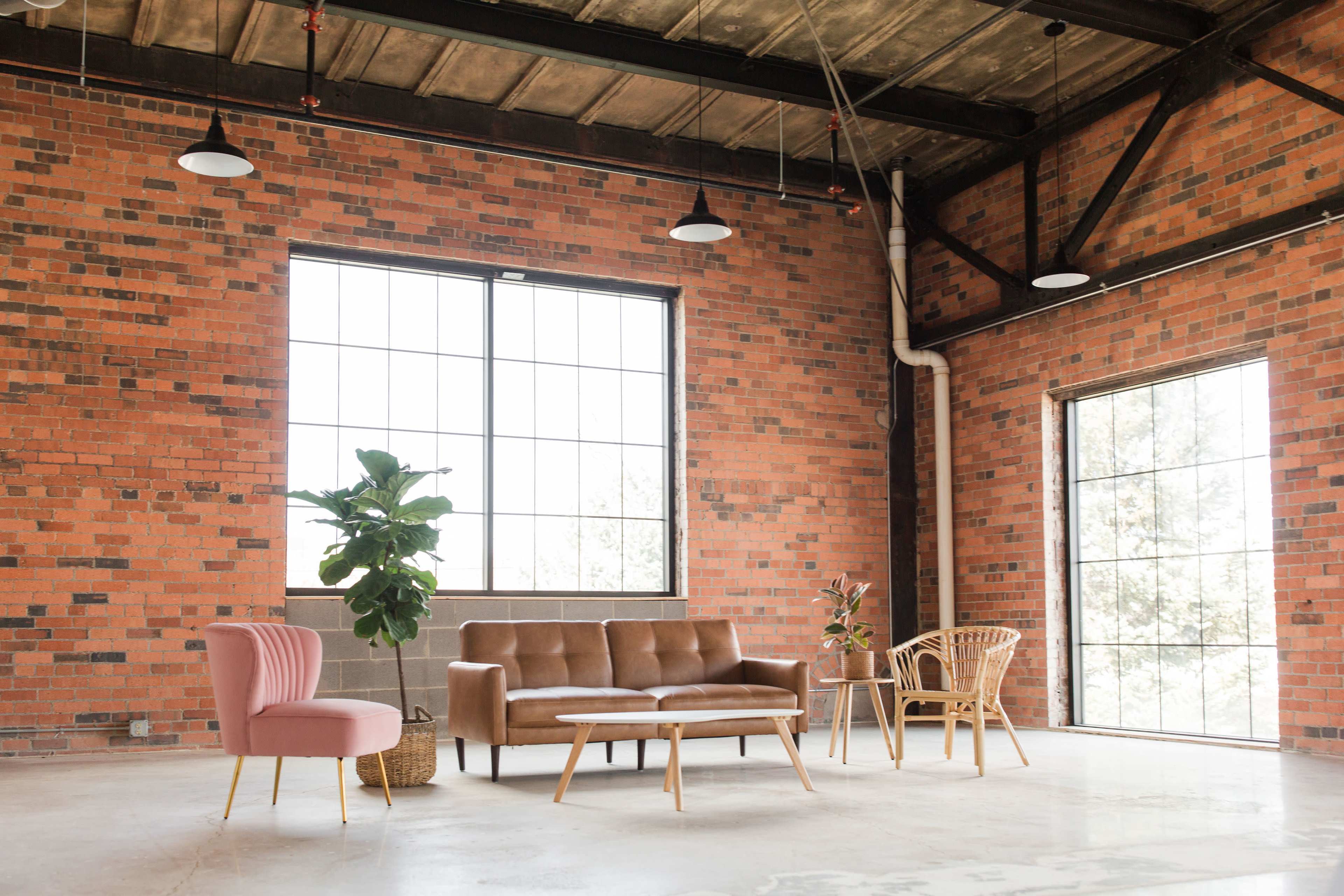 A modern interior with a brown sofa, a pink armchair, a wooden coffee table, and large windows in a space with exposed brick walls.