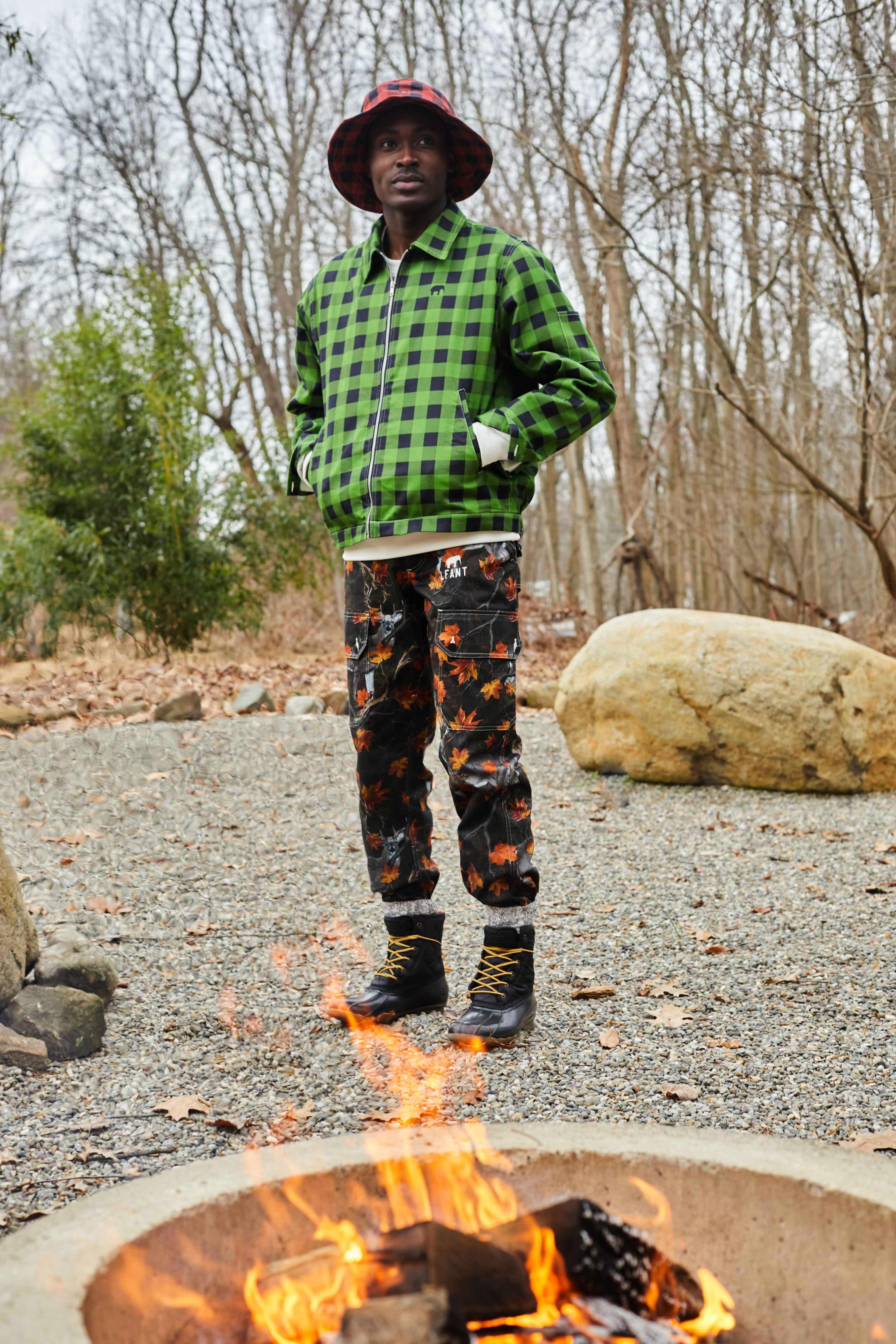 A person stands near a fire pit surrounded by stones, wearing a green and black checkered jacket and patterned pants in a natural setting with bare trees.