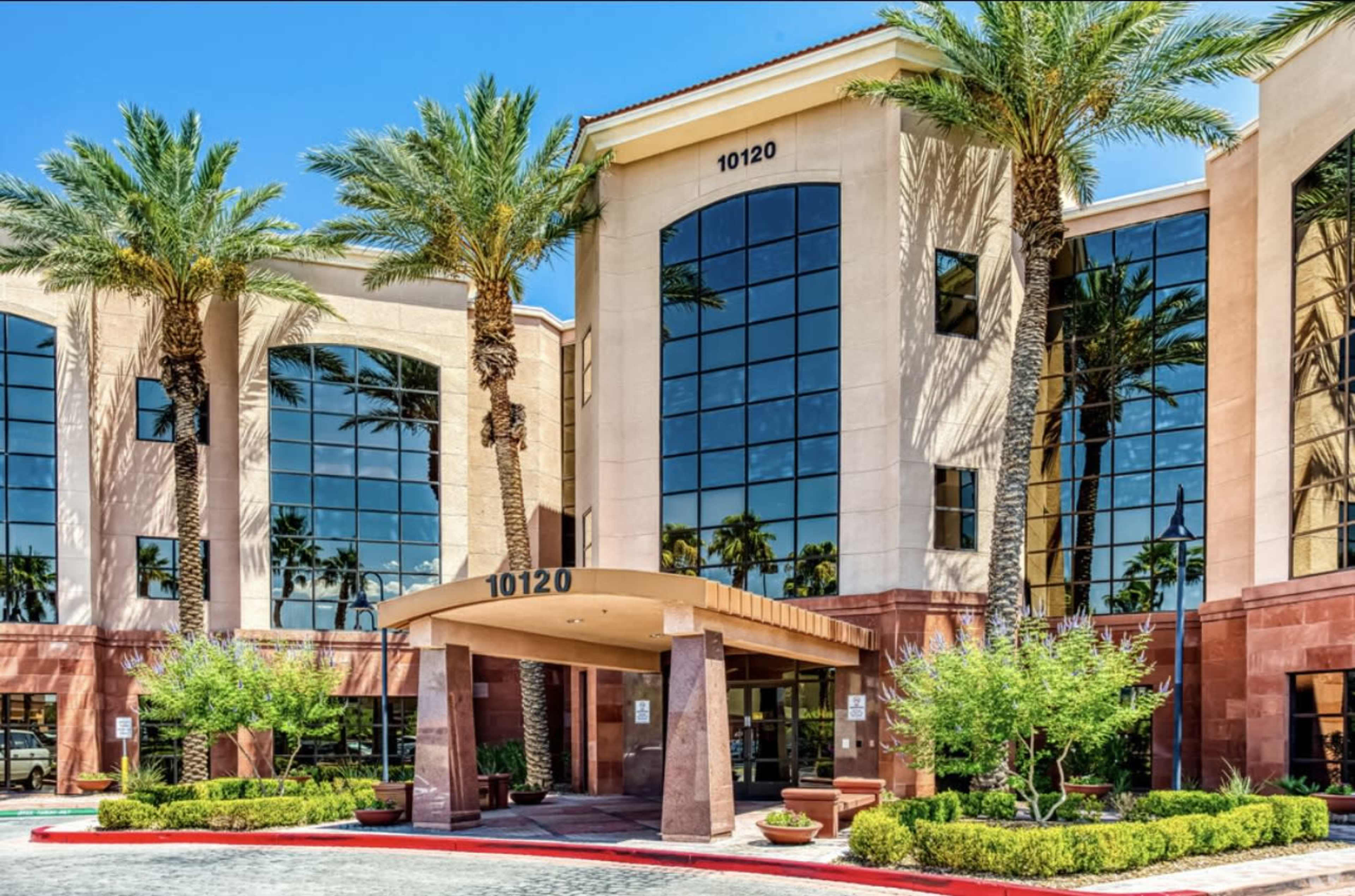 A modern office building with large glass windows, palm trees, and landscaped entryway.