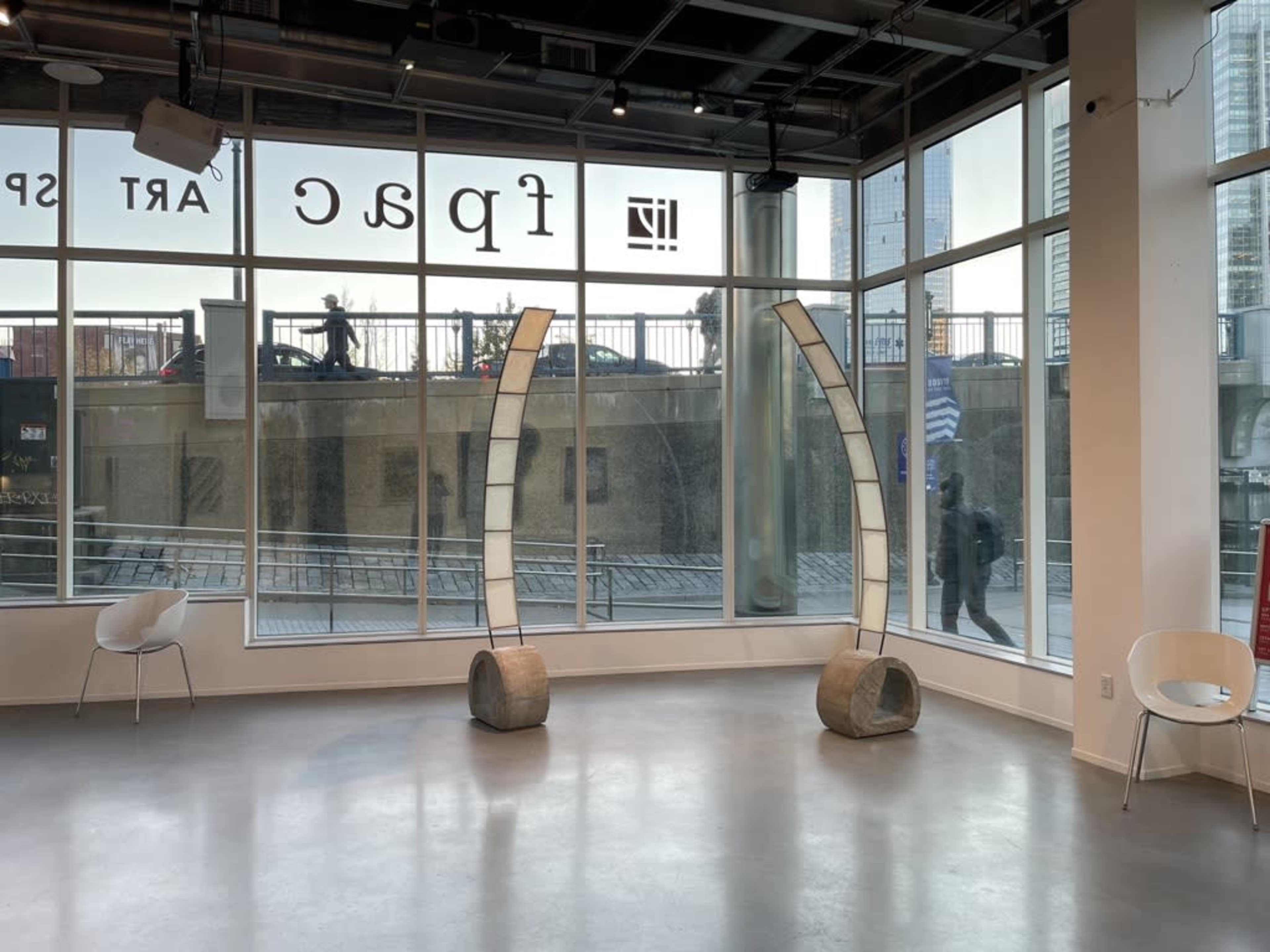 The image shows a modern art gallery space with large windows overlooking a street, featuring two abstract sculptures and minimalist seating.