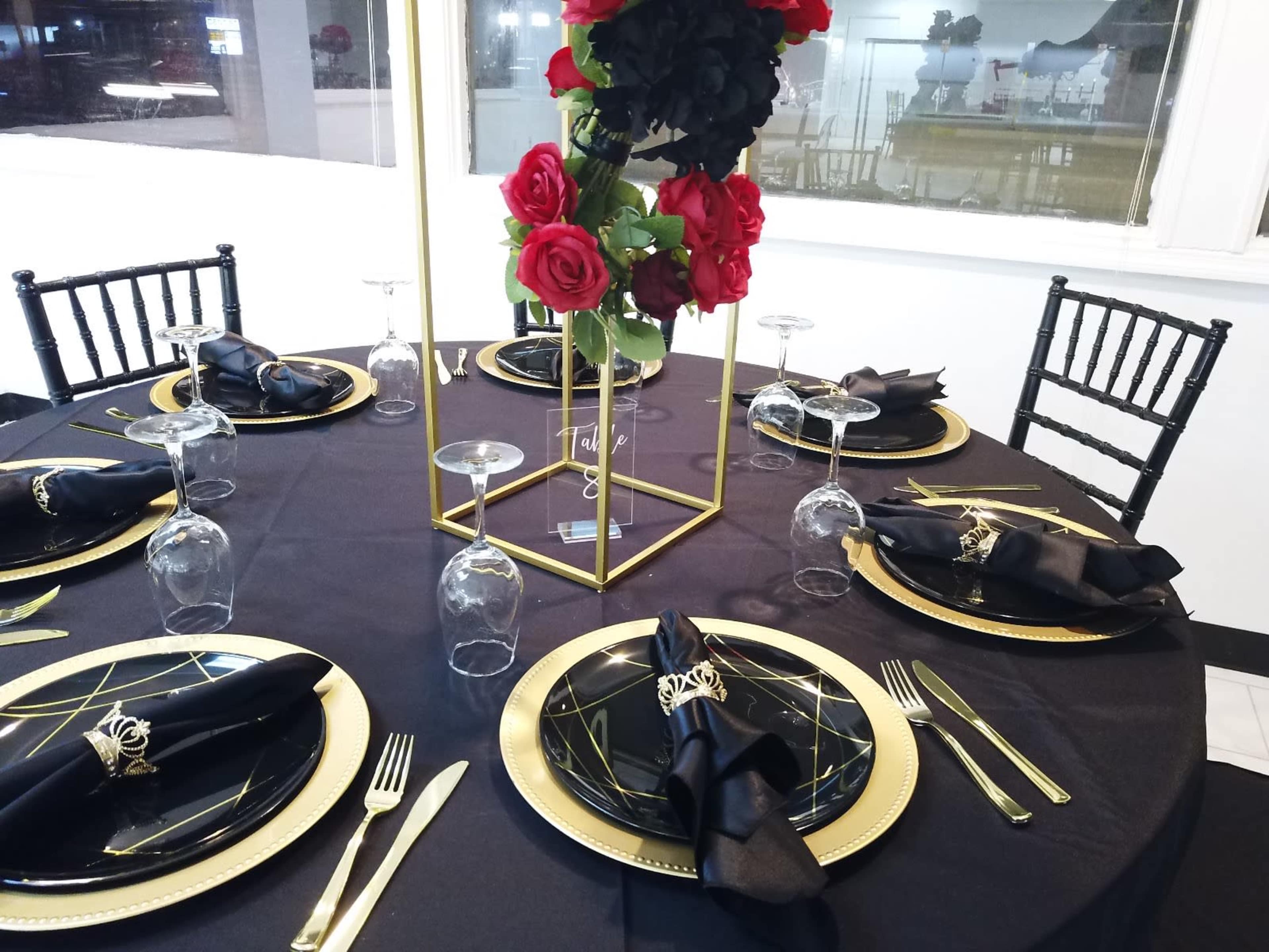 A round table is elegantly set with black napkins, golden plates, and glassware, adorned with a floral centerpiece of red roses.
