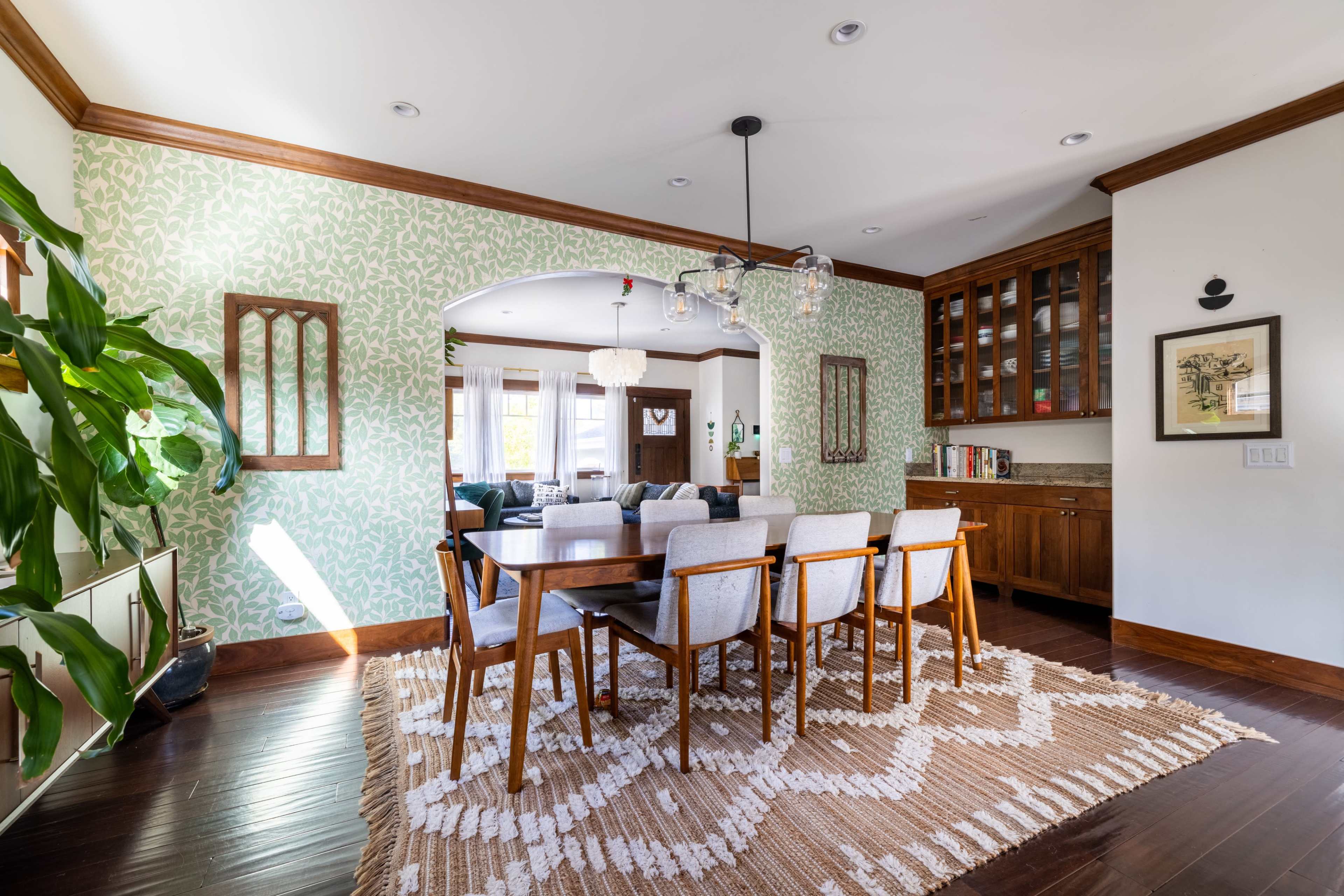 The dining area features a wooden table surrounded by upholstered chairs, with a patterned rug beneath and green wallpaper in the background.