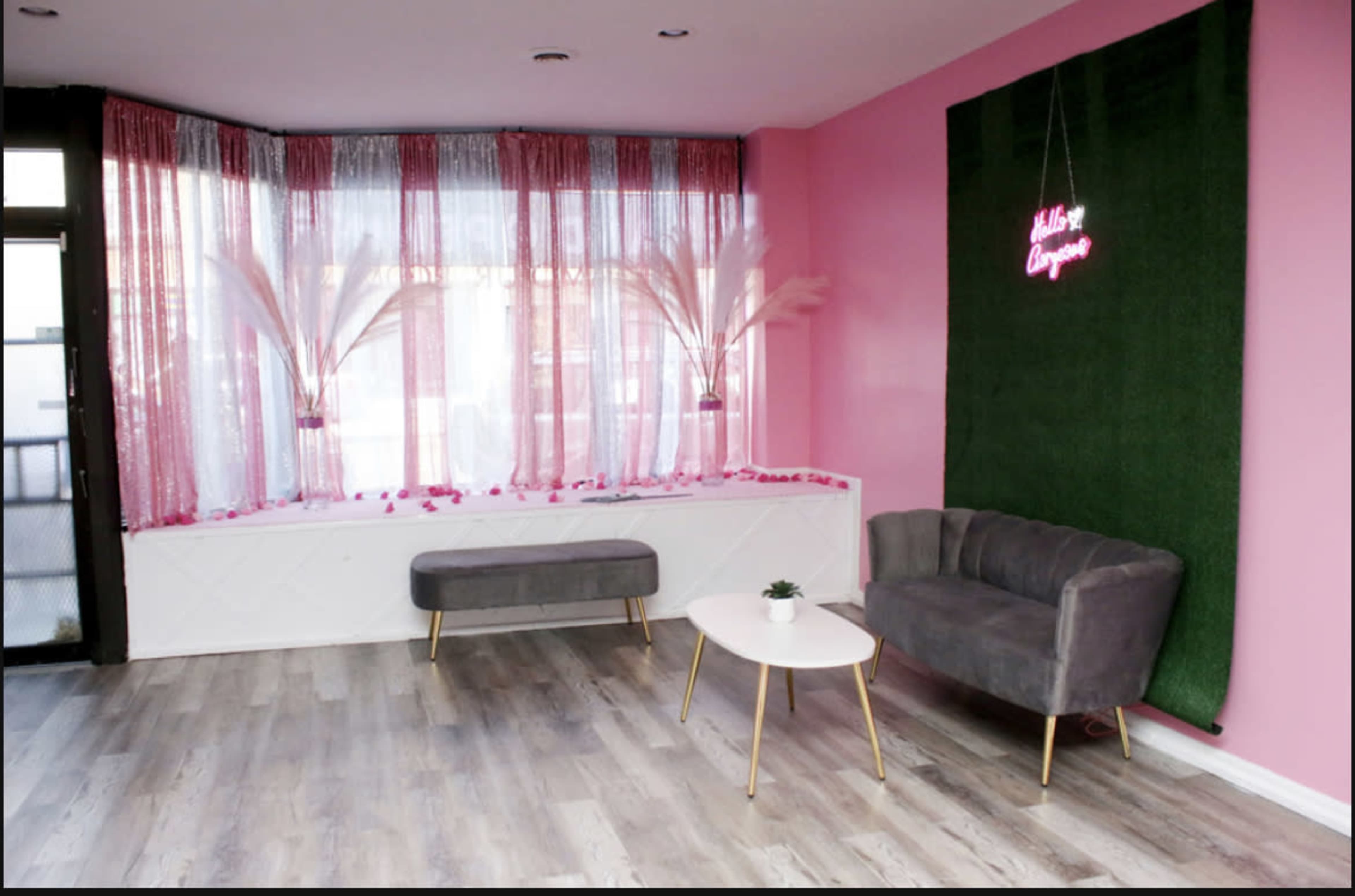 A modern indoor space with pink walls, a faux grass feature, a small table, and two grey sofas, accented by decorative plants and neon signage.