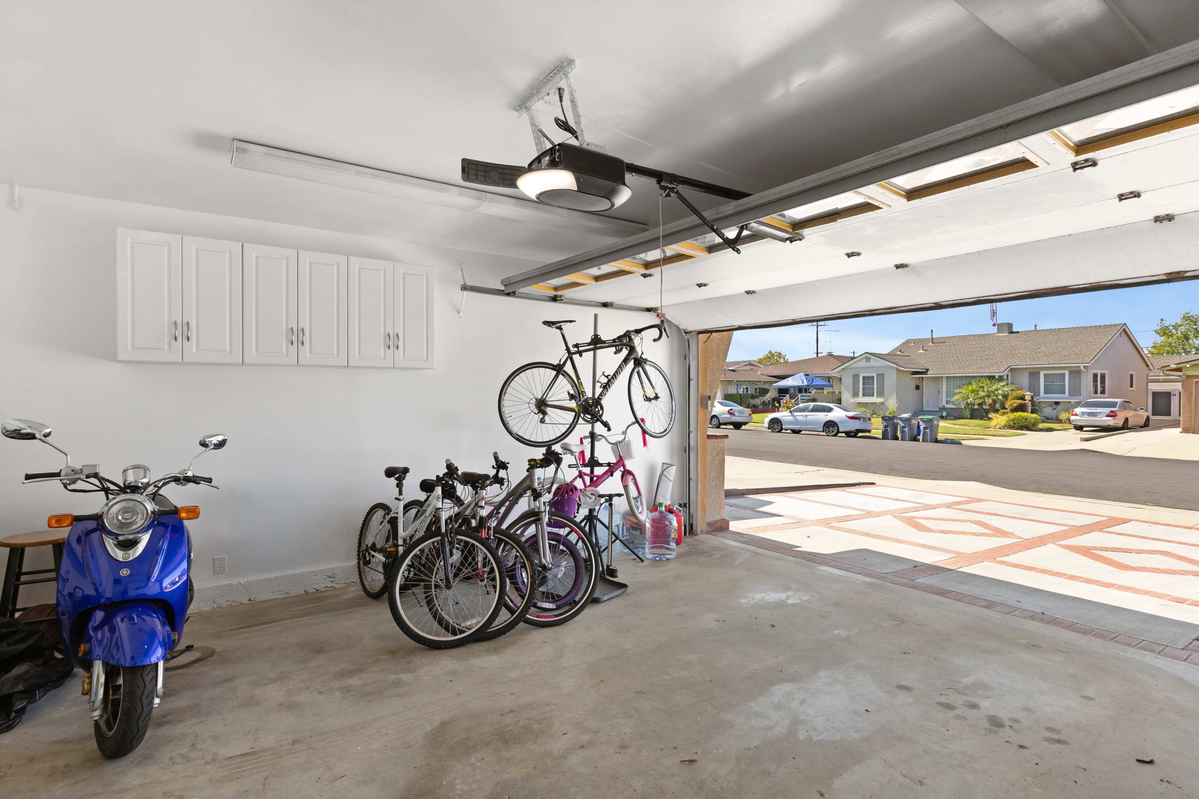 The image shows a garage with a blue scooter, several bicycles, and a door open to a residential street.