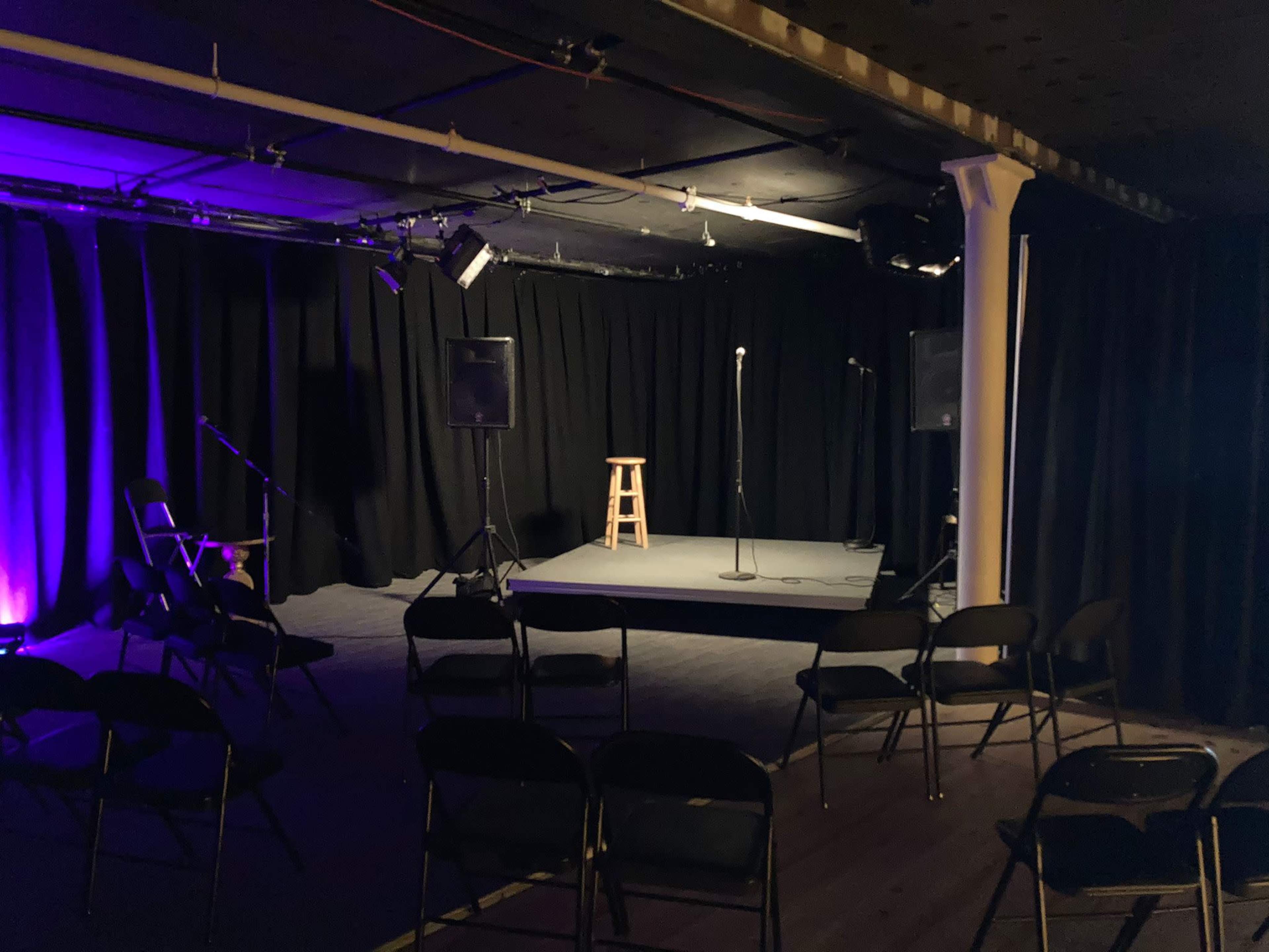 A small performance stage is set up with a microphone and a stool, surrounded by rows of empty chairs in a dimly lit room.