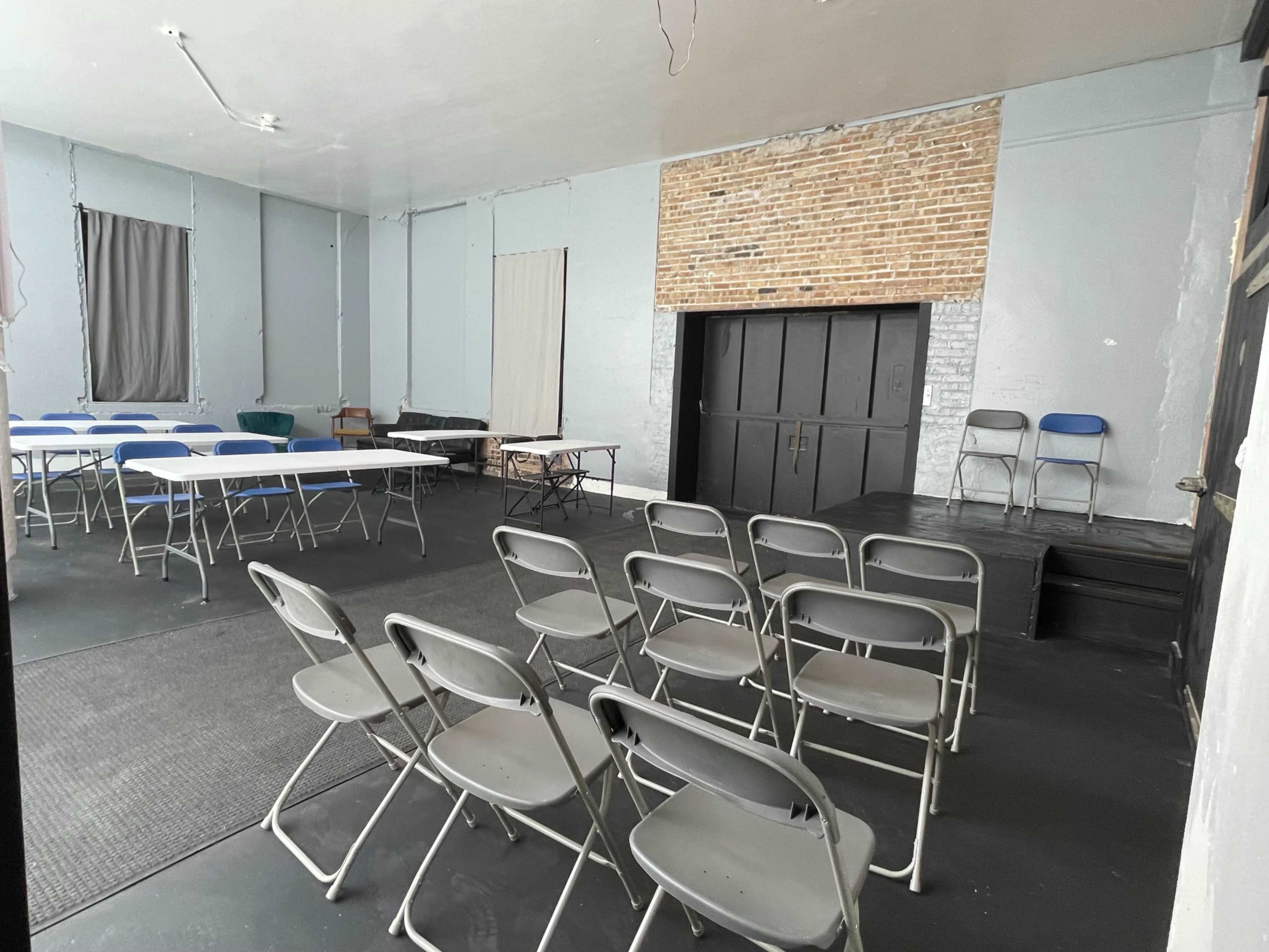 The image shows a meeting room with several rows of folding chairs, tables set up for an event, and a brick wall section in the back.