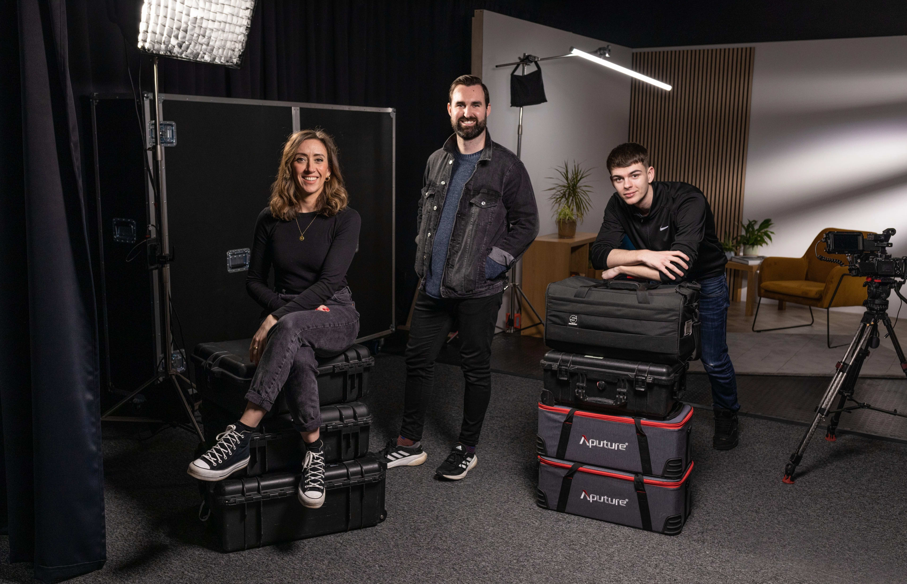 Three individuals pose on stacked equipment cases in a studio setting, with cameras and lighting gear visible in the background.