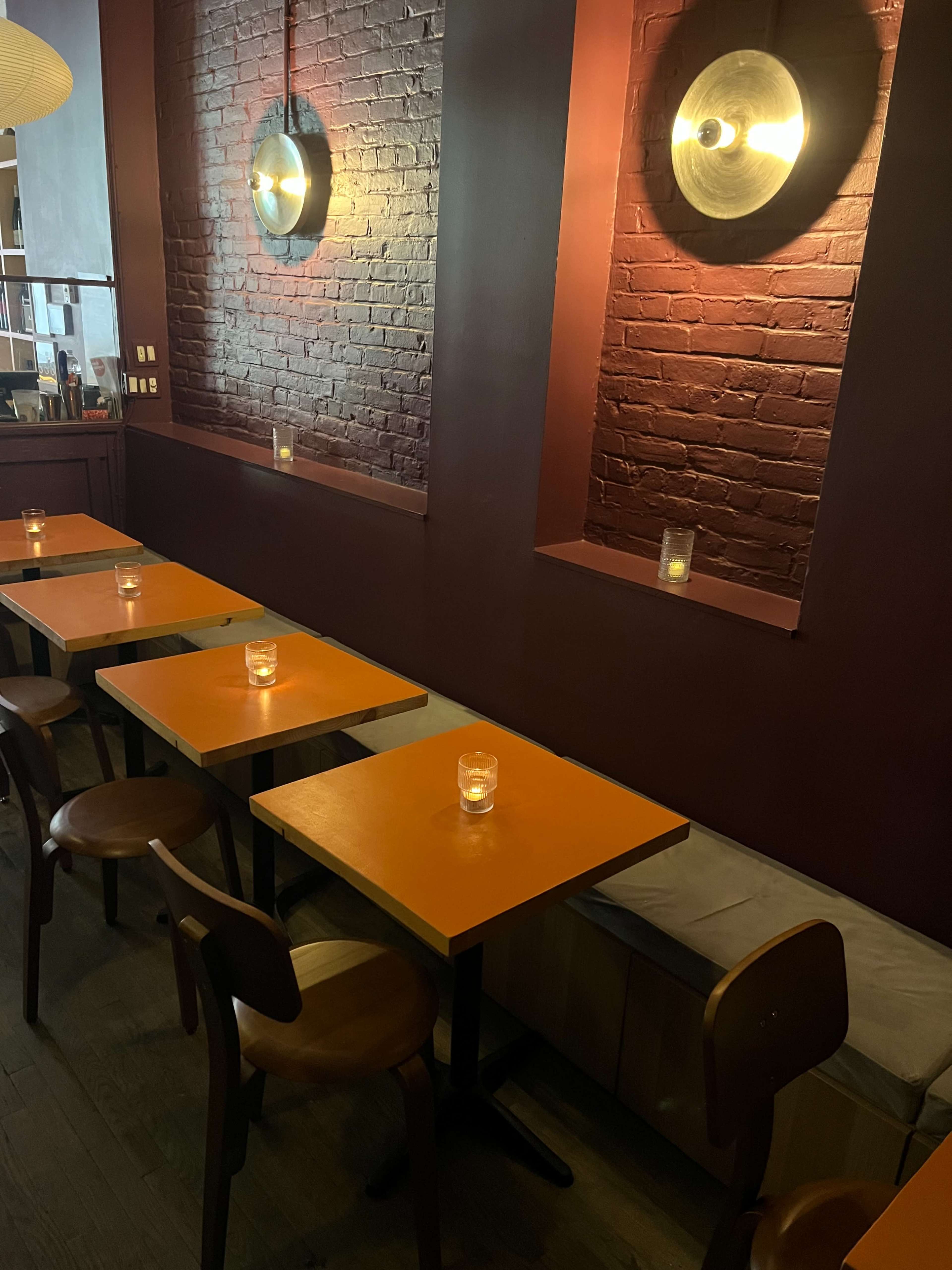 The image depicts a cozy restaurant interior with orange tables, wooden chairs, and soft lighting from wall-mounted fixtures.