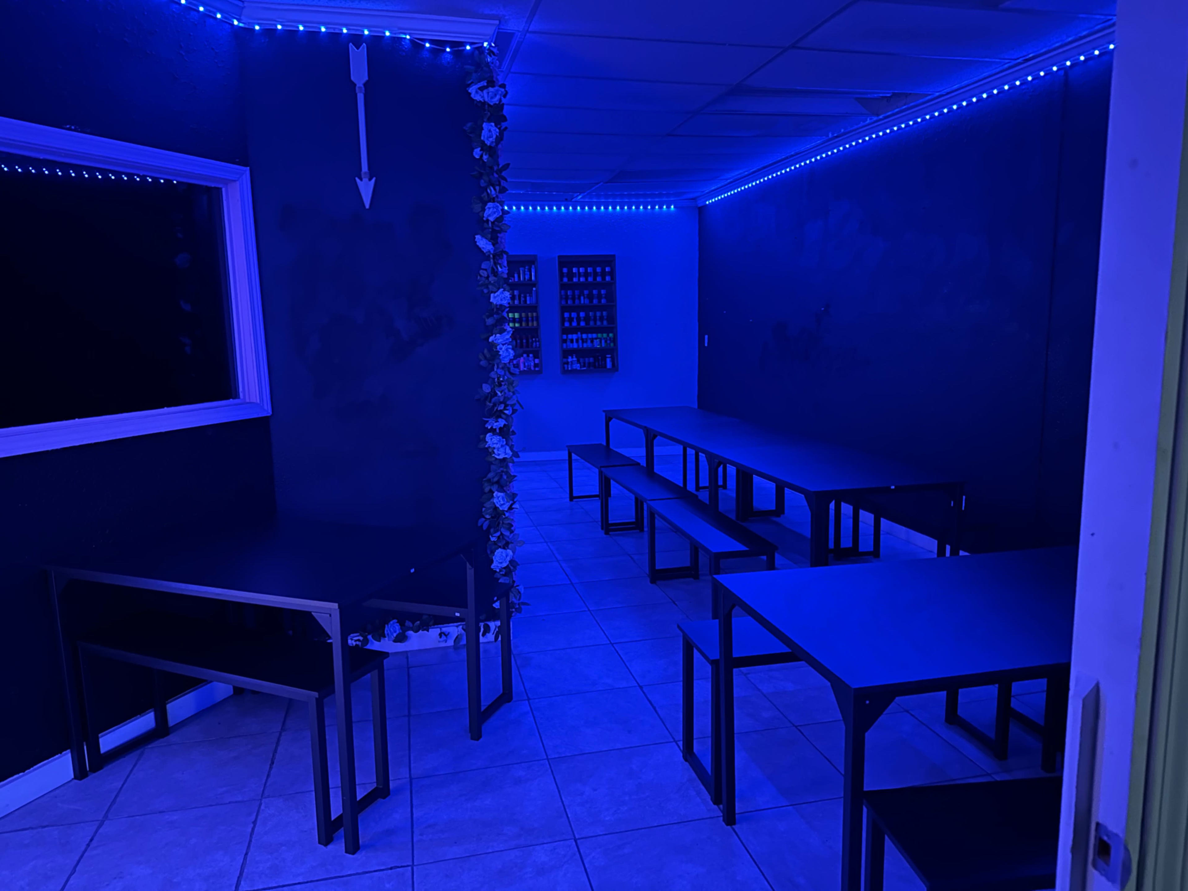 A dimly lit room with blue lighting, featuring two rows of metal tables and a wall adorned with bottles.
