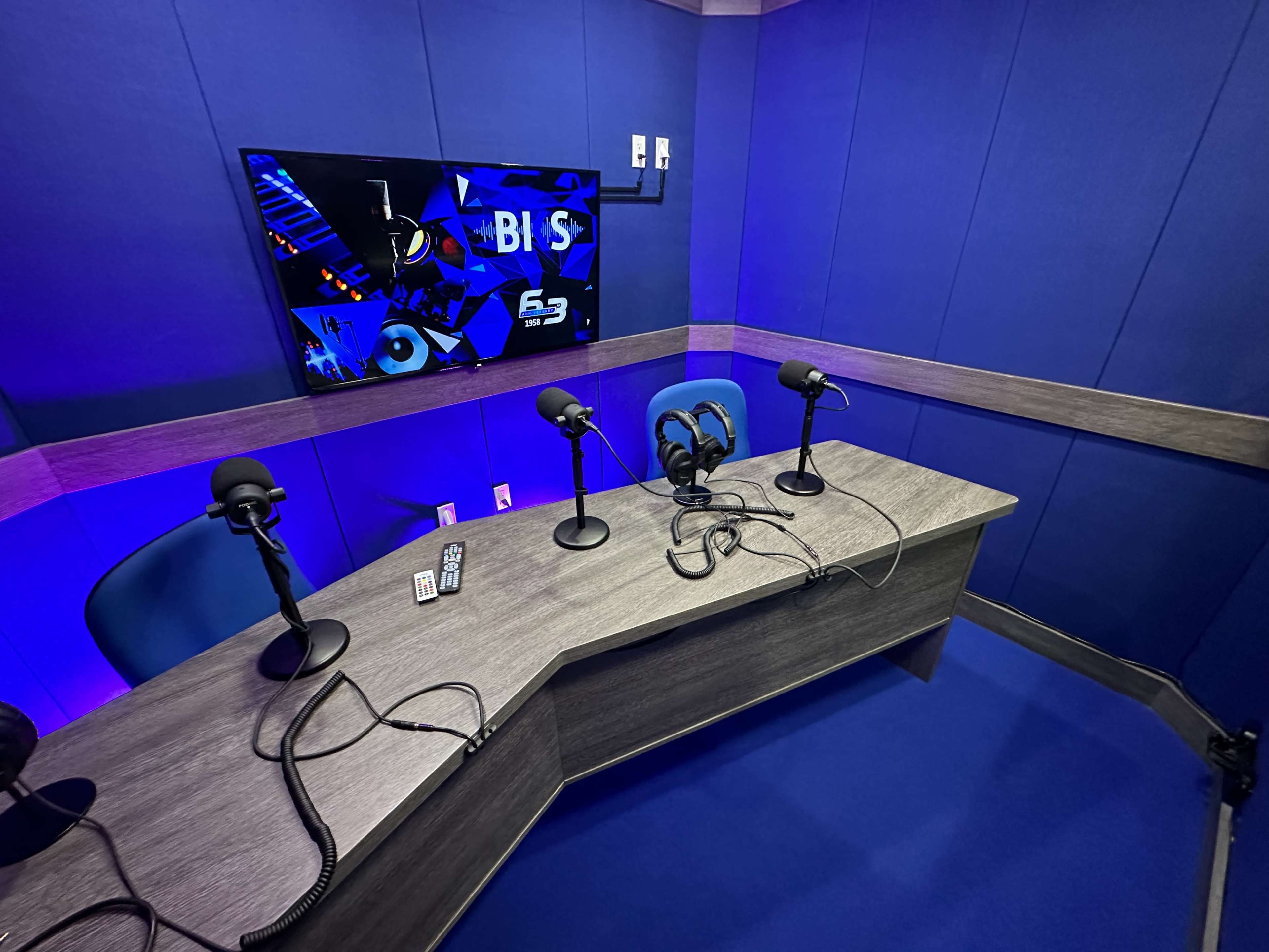 A modern recording studio with a blue backdrop, featuring a wooden desk and three microphones on stands.