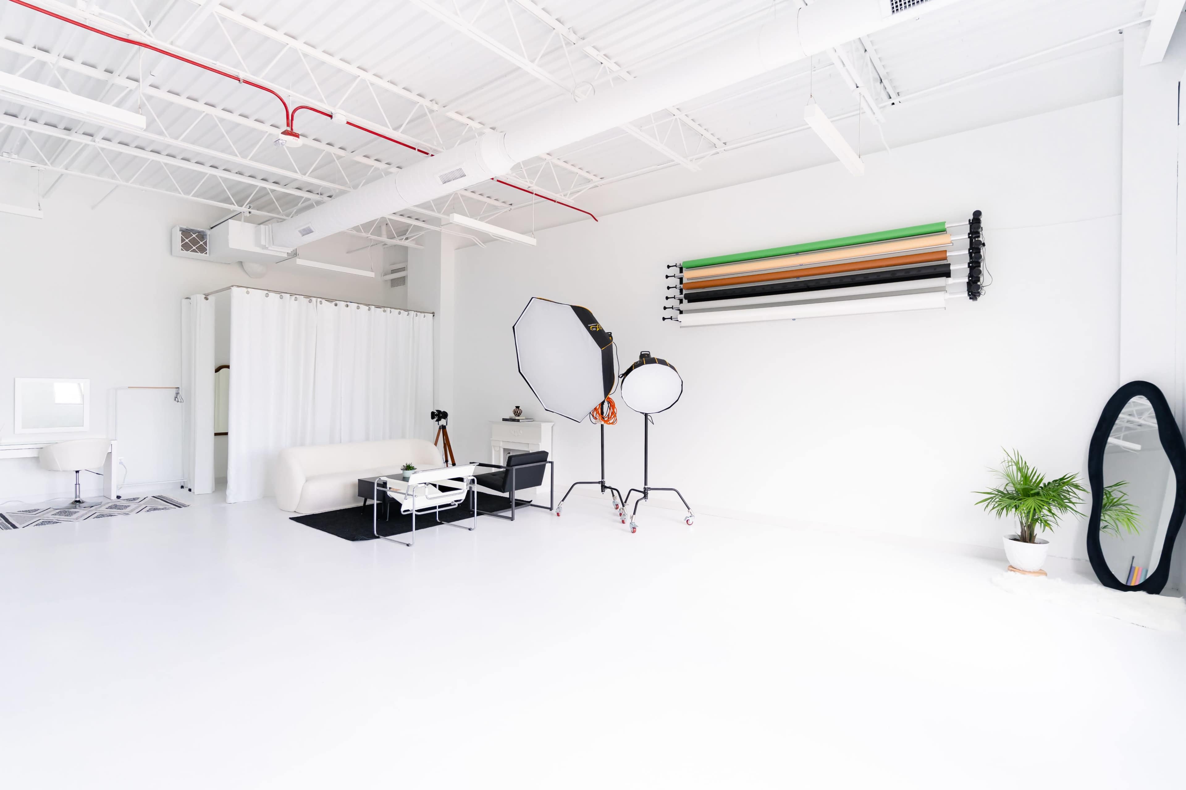 A bright, minimalist studio space with a white floor, a black couch, photography equipment, and a large mirror.