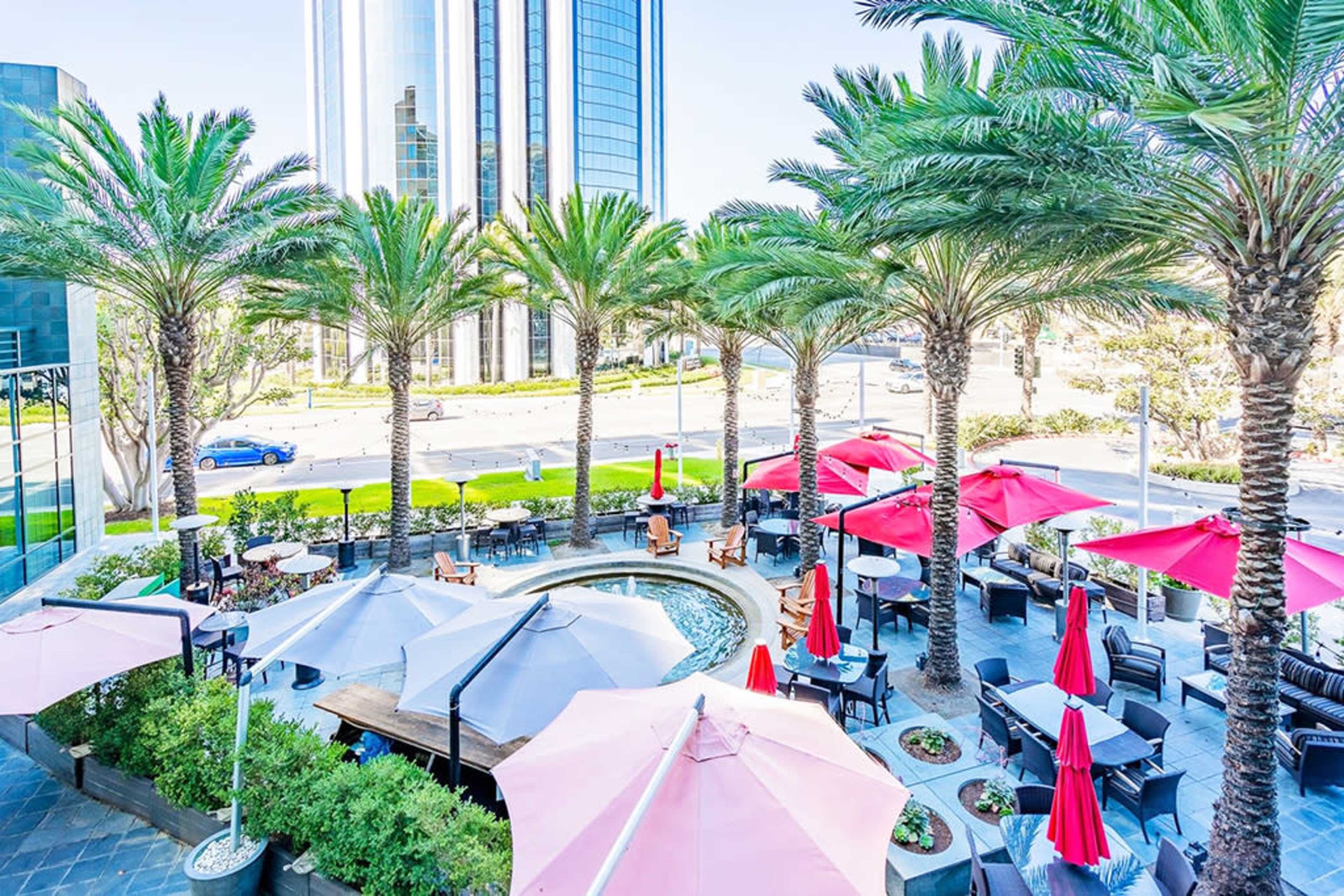 The image shows an outdoor dining area with palm trees and red umbrellas surrounding a small water feature, set against a backdrop of tall modern buildings.