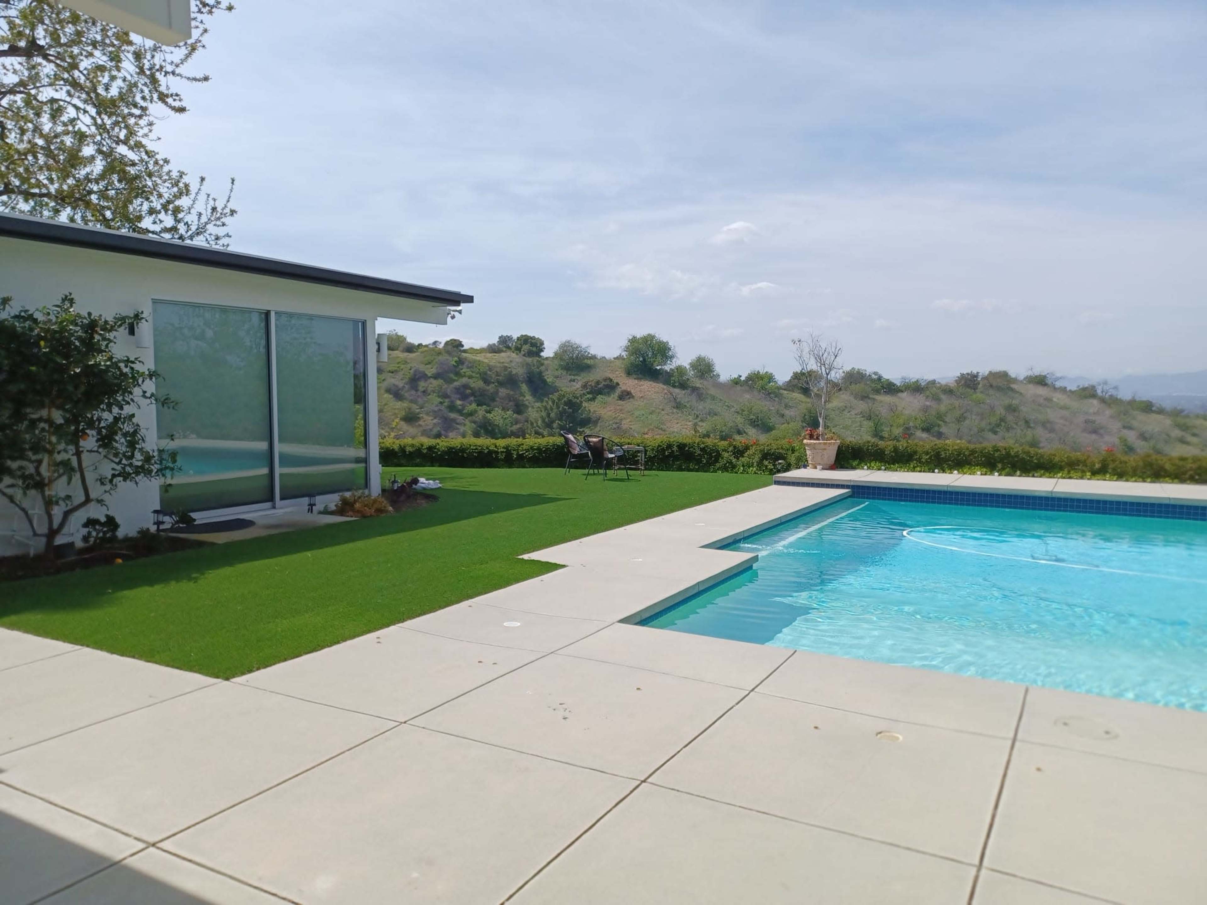 A modern backyard features a swimming pool, a patio with concrete tiles, and a grassy area adjacent to a hillside.