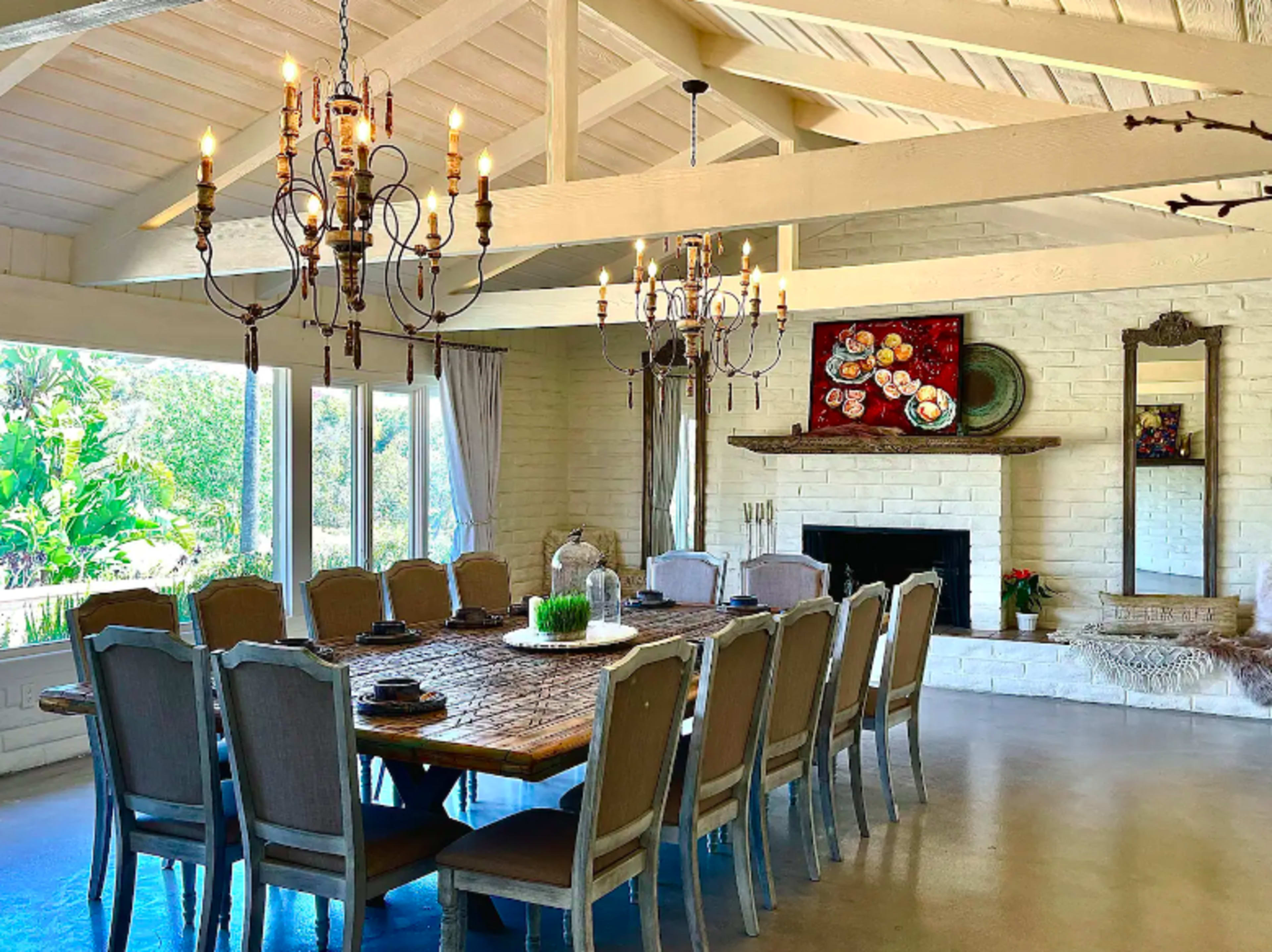 A spacious dining area features a long wooden table surrounded by upholstered chairs, with large windows allowing natural light and a fireplace accenting the room.