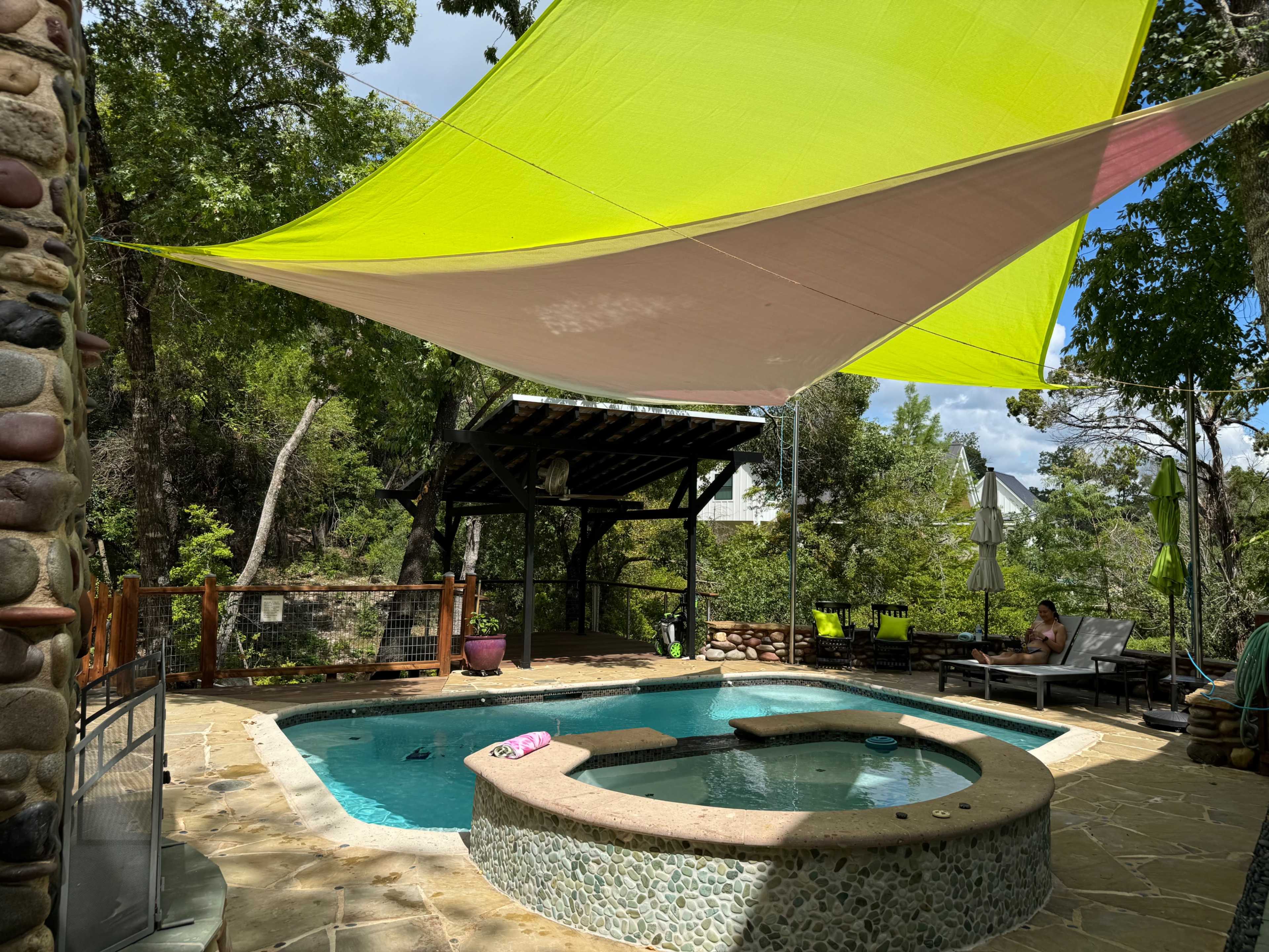 A bright green and pink shade sail covers a swimming pool area with a hot tub, surrounded by trees and outdoor furniture.