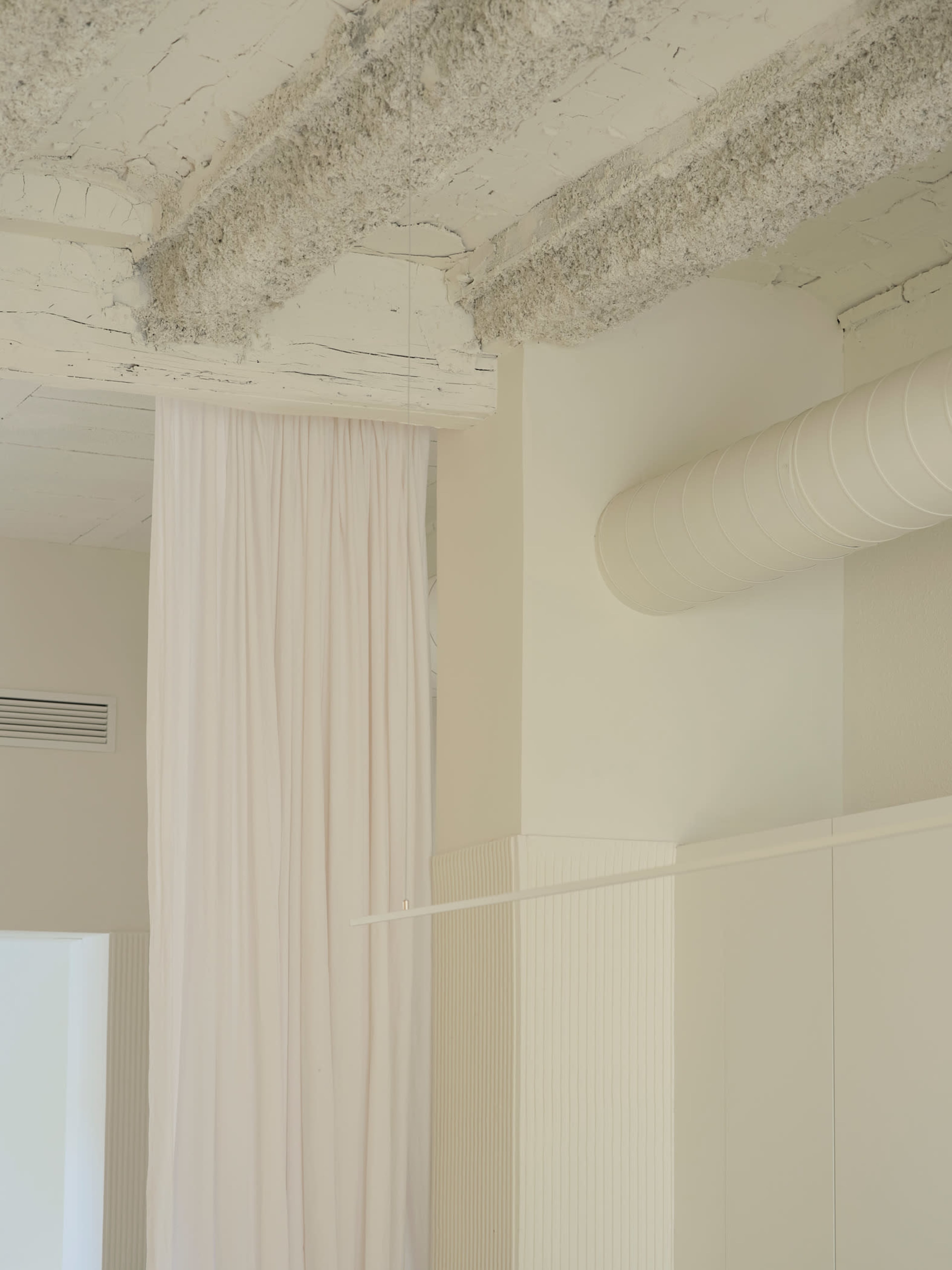The image shows a minimalist interior featuring a textured ceiling, a draped curtain, and a cylindrical ventilation duct against a neutral backdrop.