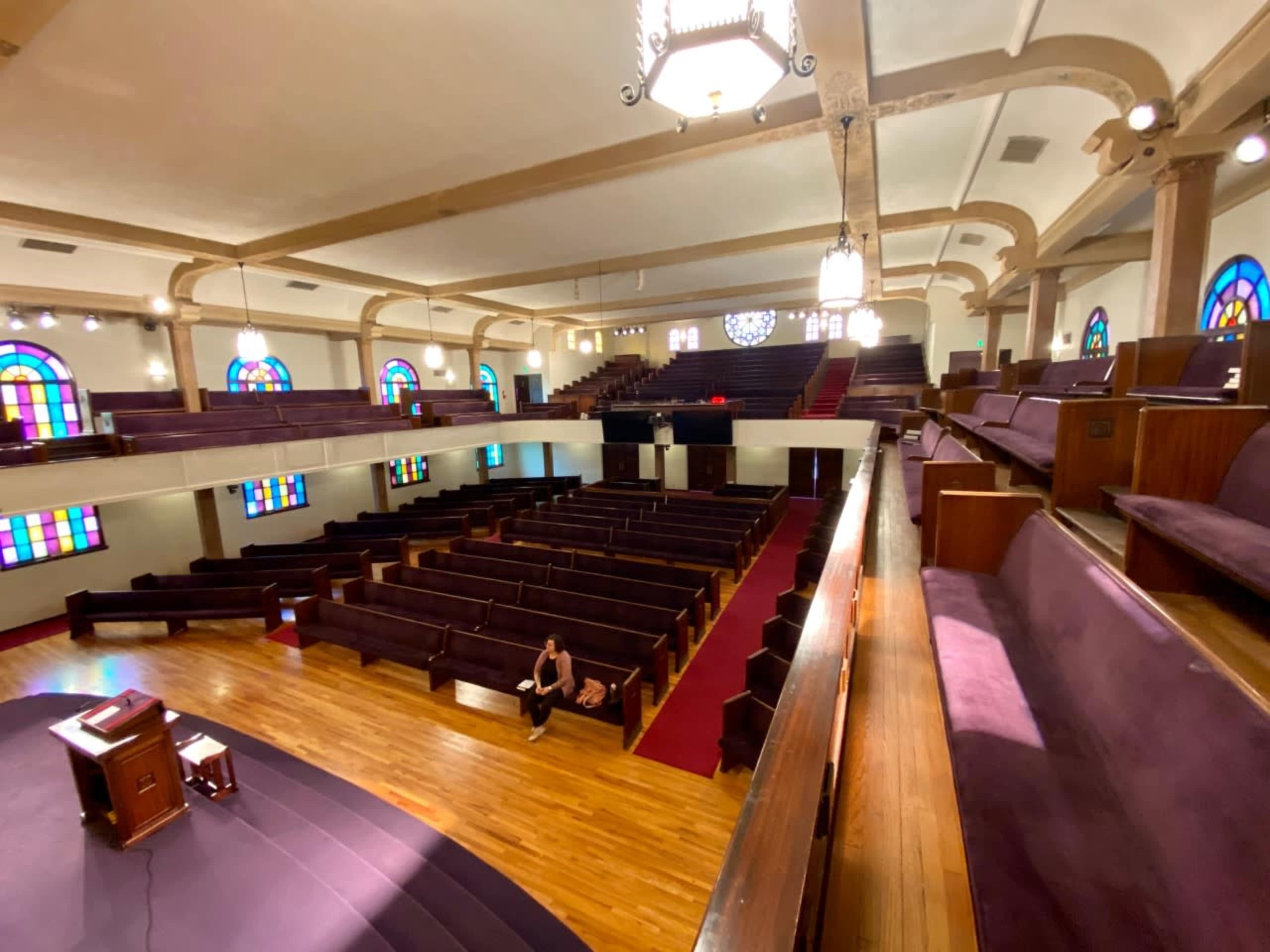 An empty church interior with wooden pews, stained glass windows, and a wooden pulpit on a raised stage.