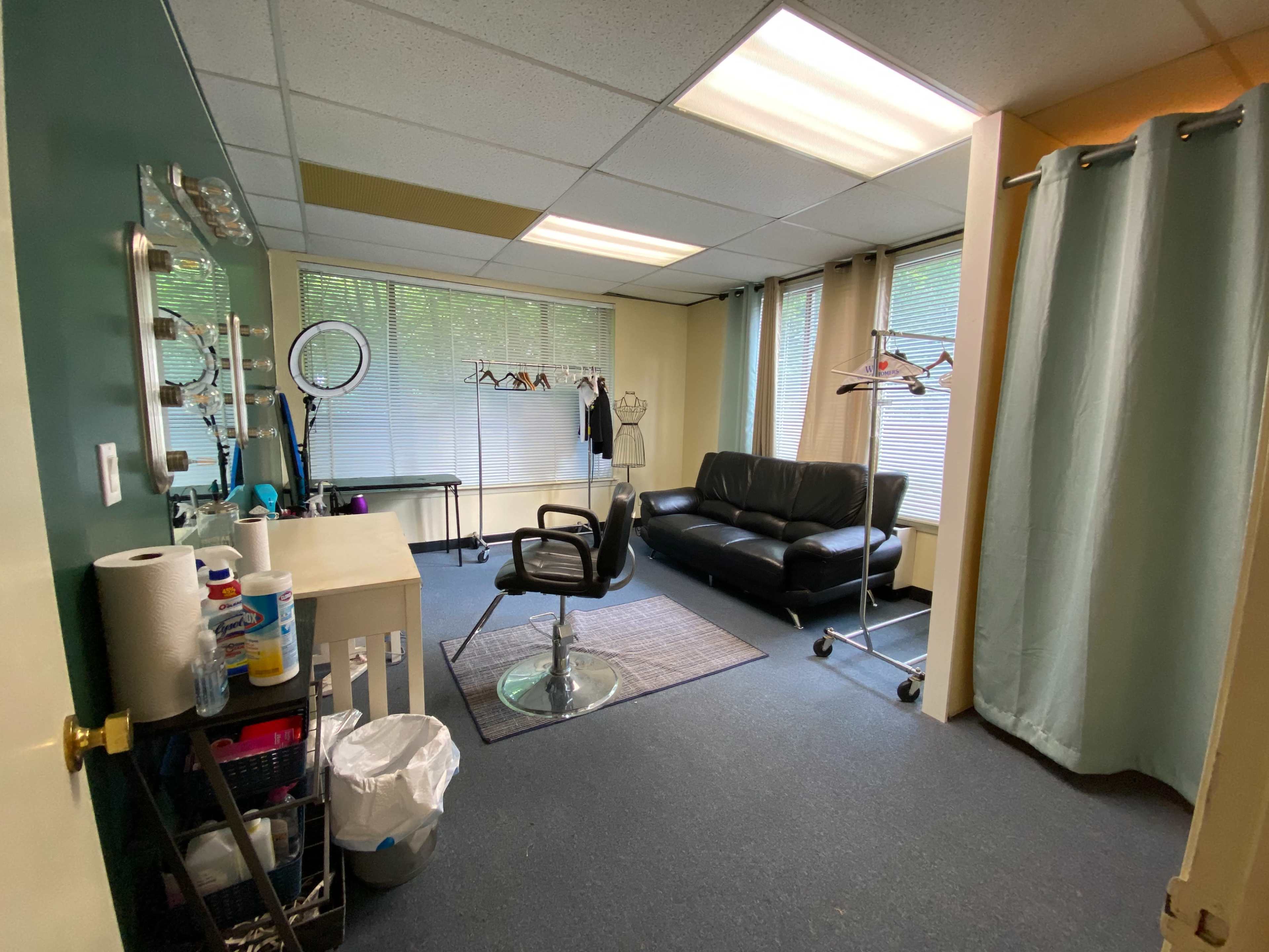 The image shows a room with a black leather sofa, a styling chair with a mirrored vanity, and clothing displayed on hangers near the windows.