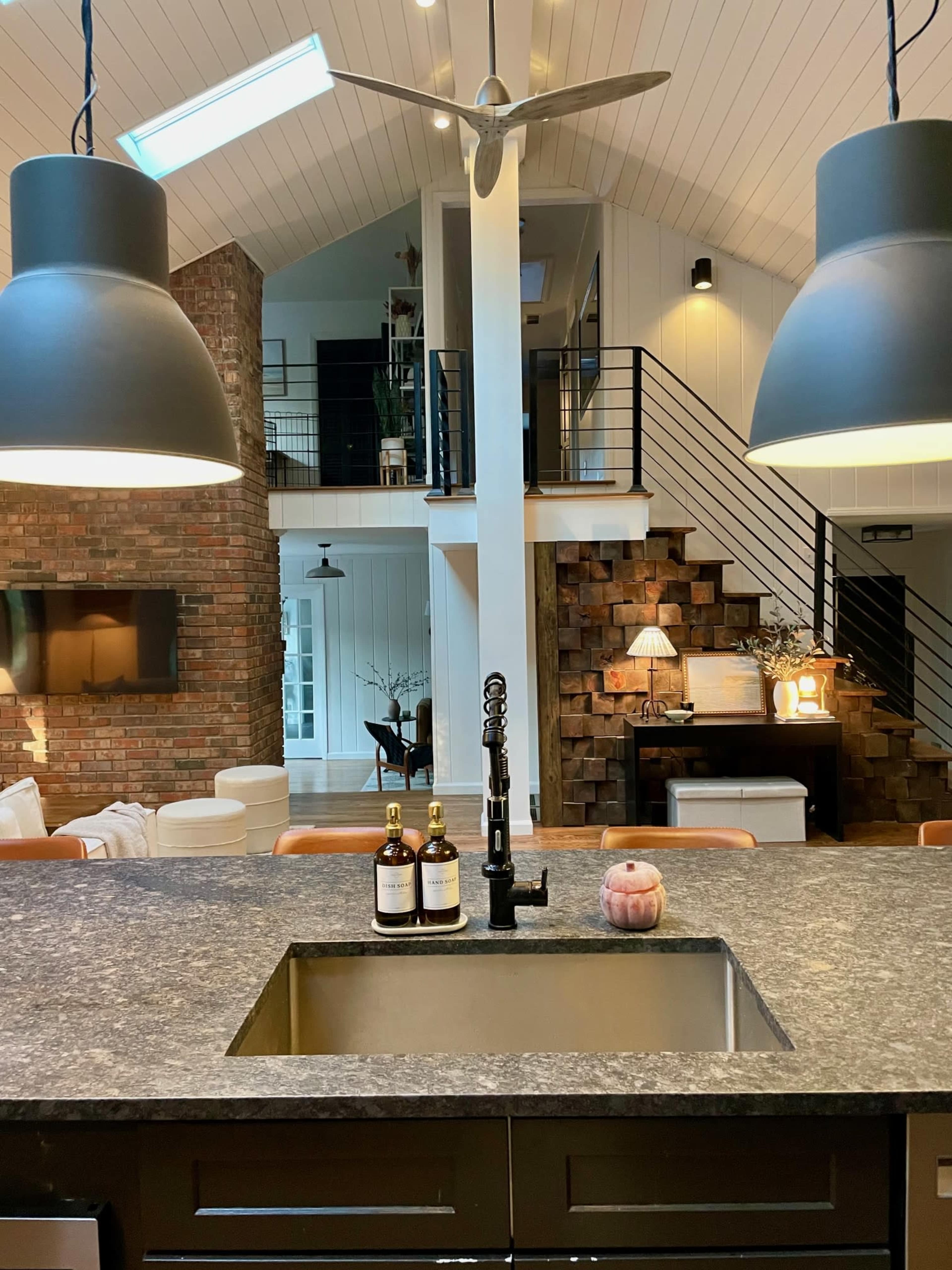 The image shows a modern kitchen with a central island featuring a sink and illuminated by two large pendant lights, alongside a staircase with a brick wall in the background.