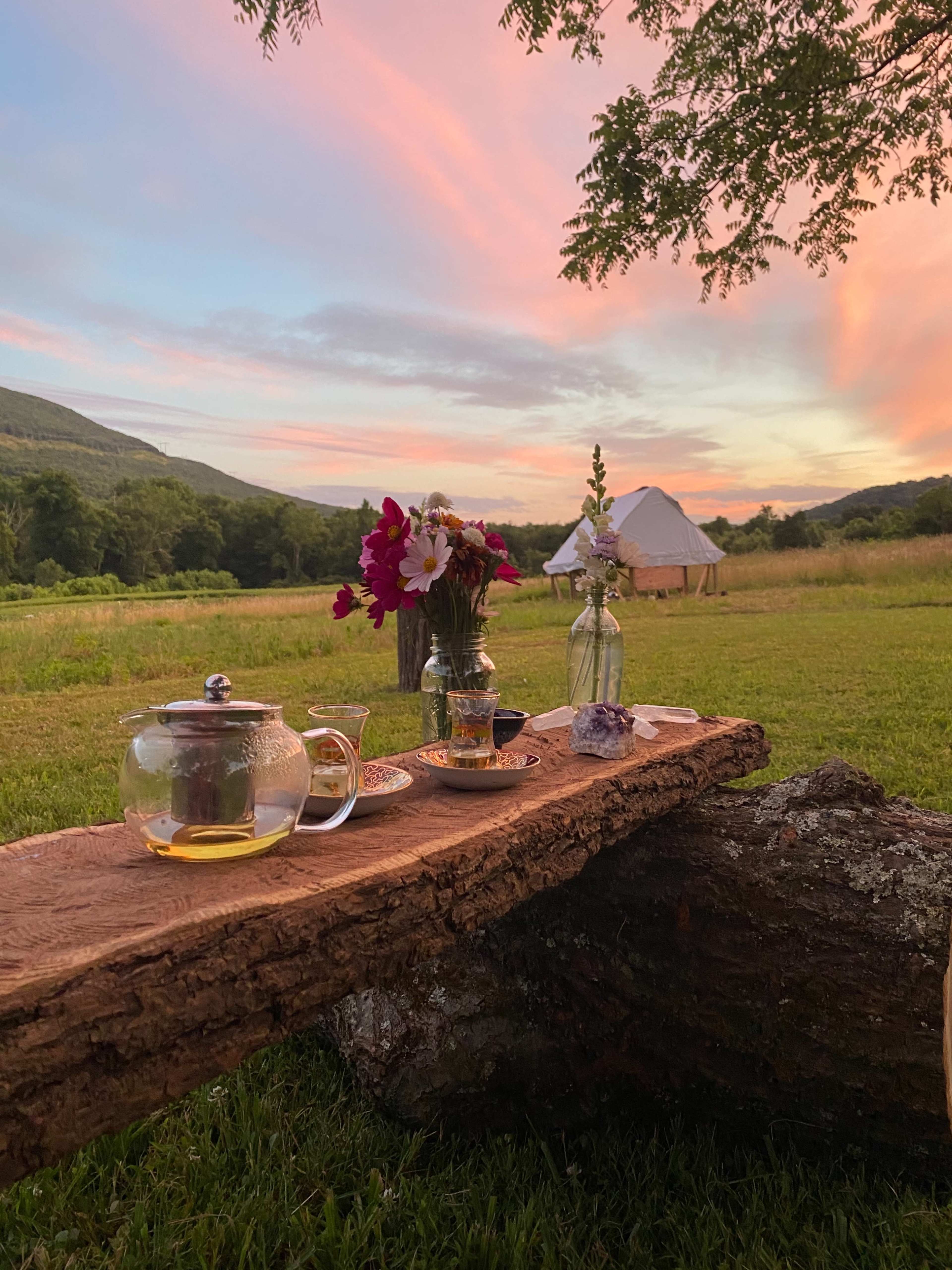 A wooden table is set with a teapot, cups, and flowers, with a tent in the background under a colorful sunset sky.