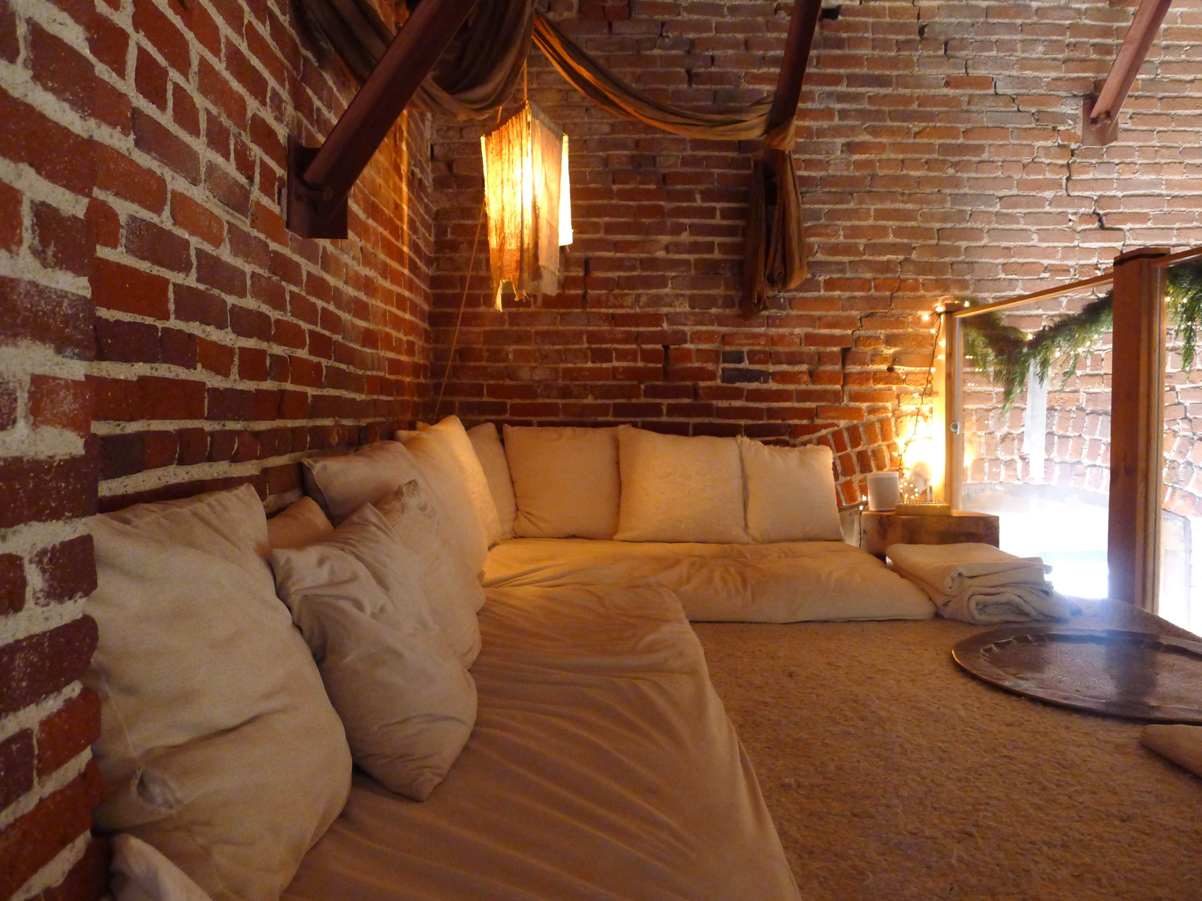 The image shows a cozy seating area with a sofa covered in beige cushions, surrounded by exposed brick walls and soft lighting.