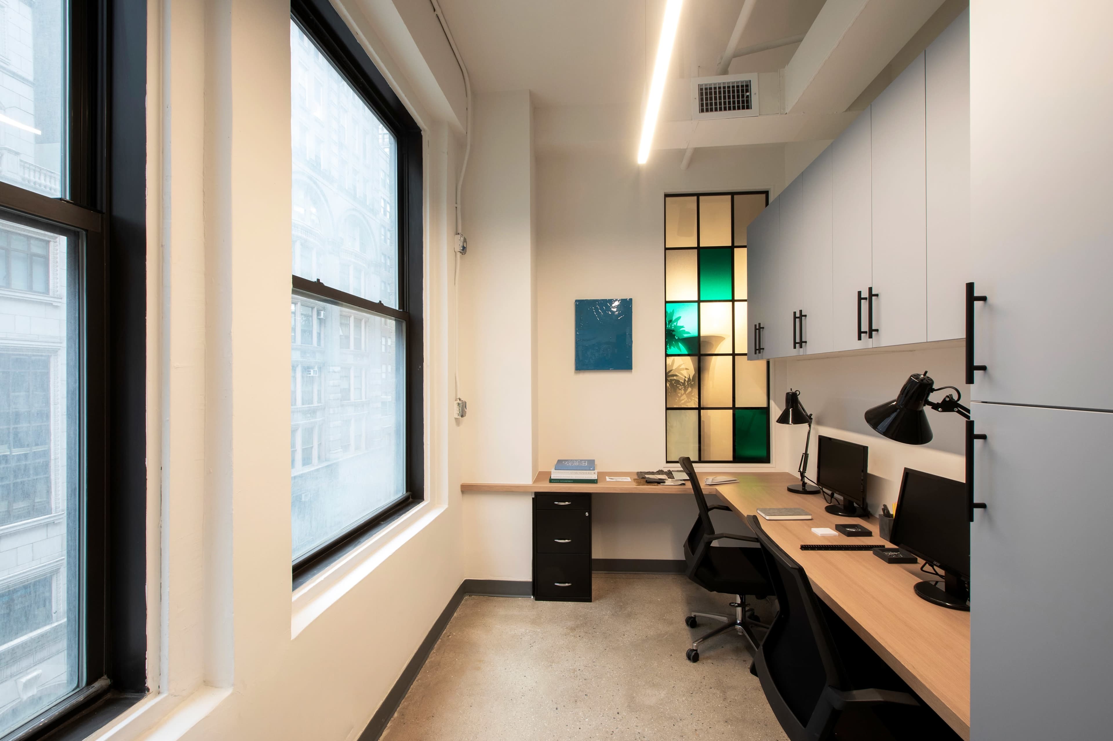 A modern office space with two desks, each equipped with a computer, beside large windows and a colorful glass panel.