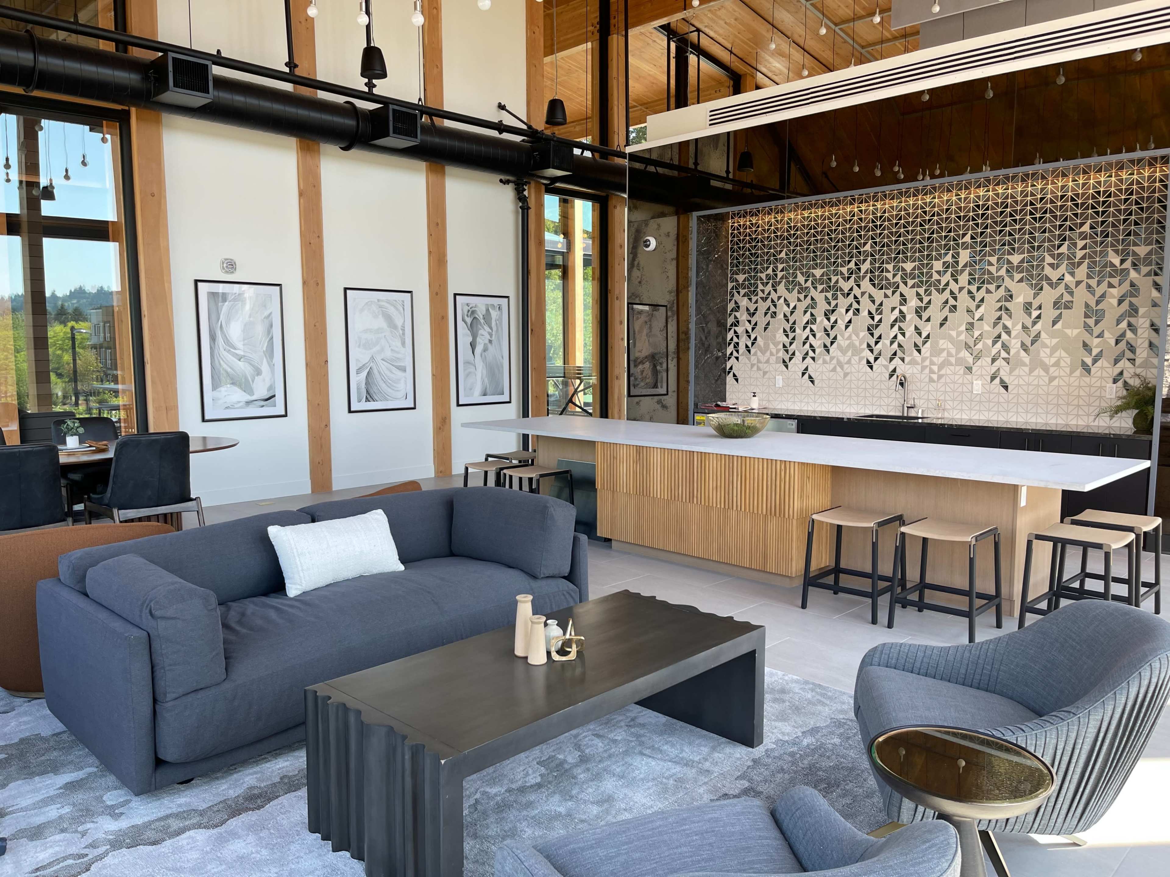 A modern living space featuring a gray sofa, a coffee table, and a kitchenette with a patterned backsplash, surrounded by large windows.