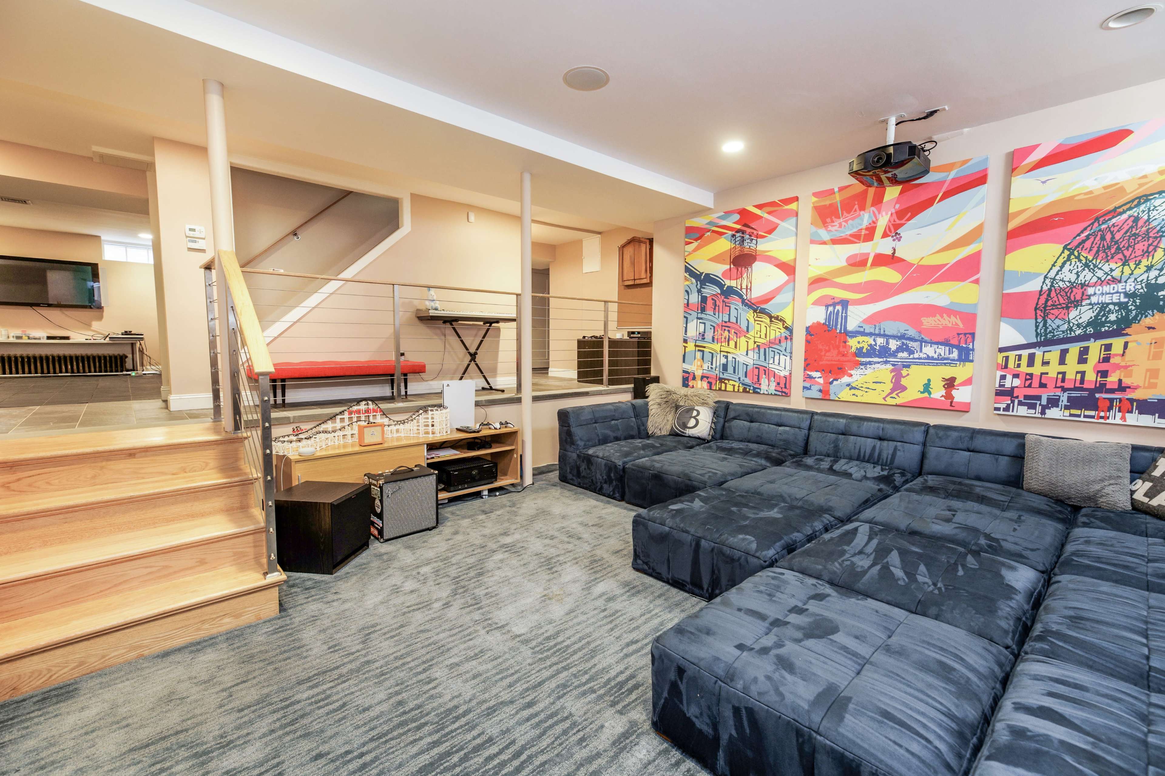 A spacious basement lounge features a large blue sectional sofa, a colorful mural on the wall, and a staircase leading to an upper level.