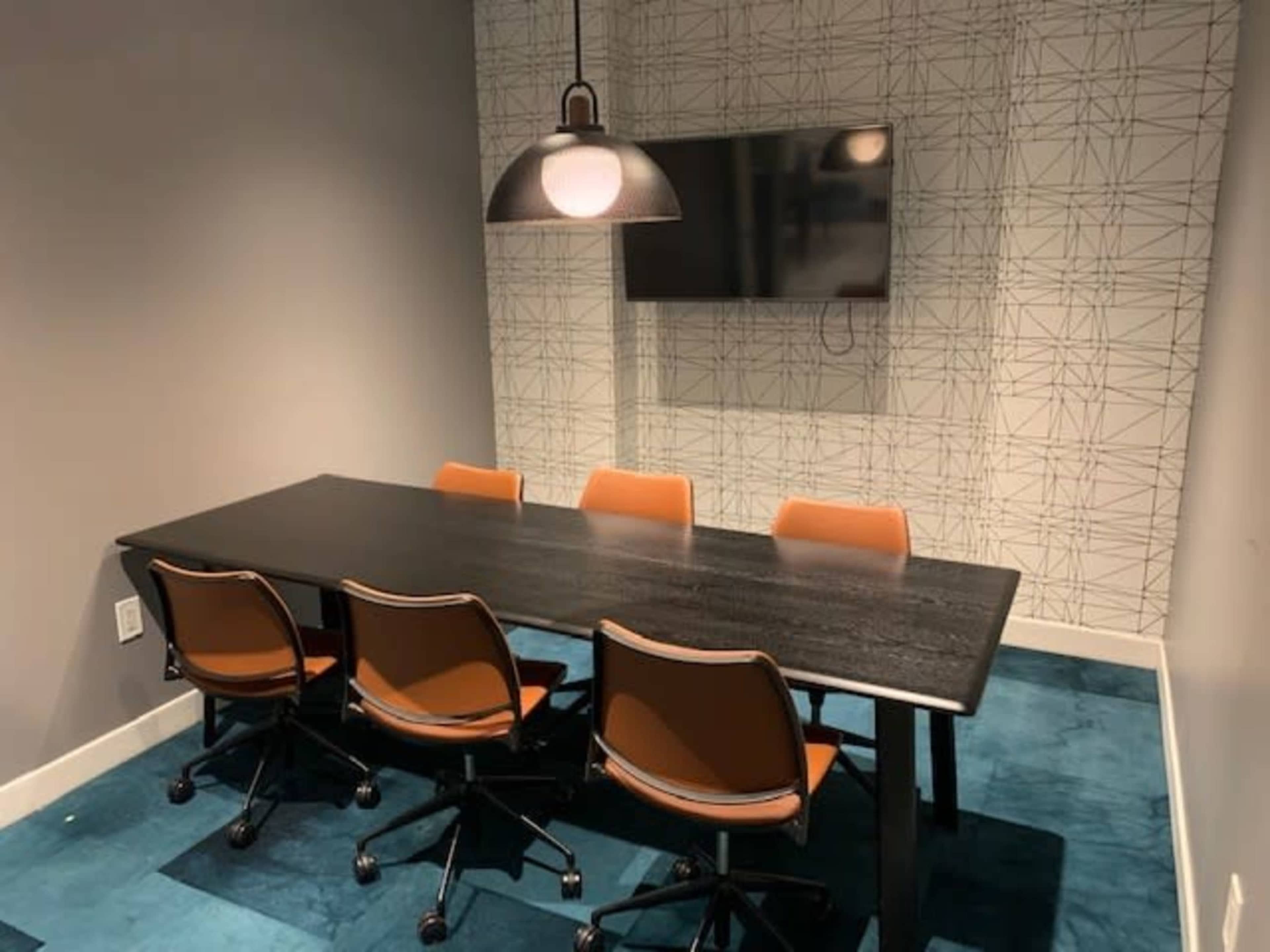 A modern conference room features a long black table surrounded by five orange chairs, with a wall-mounted television and a geometric patterned backdrop.