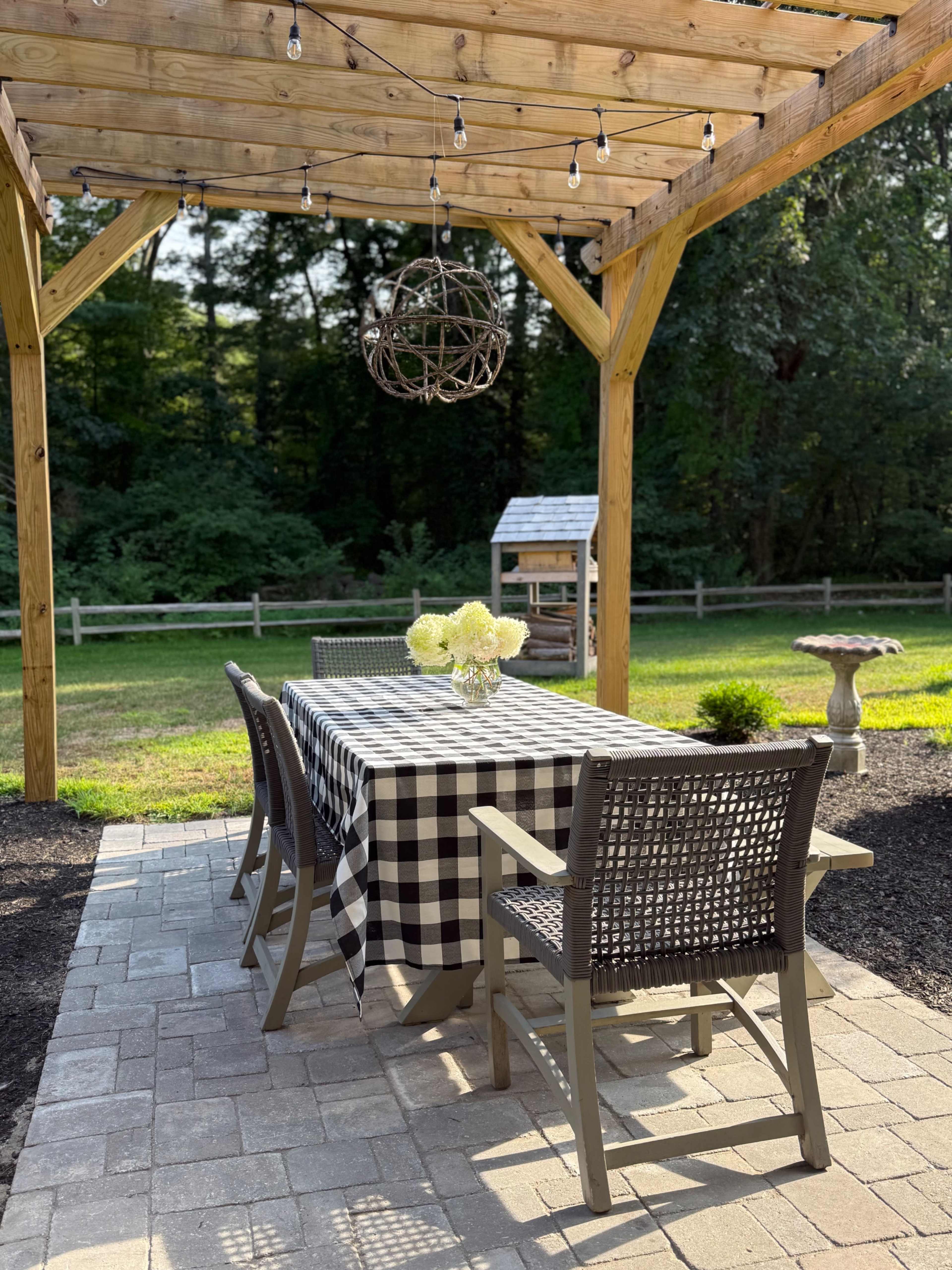 A wooden pergola with string lights shelters a checkered table set with a bouquet of flowers, surrounded by wicker chairs on a paved patio.
