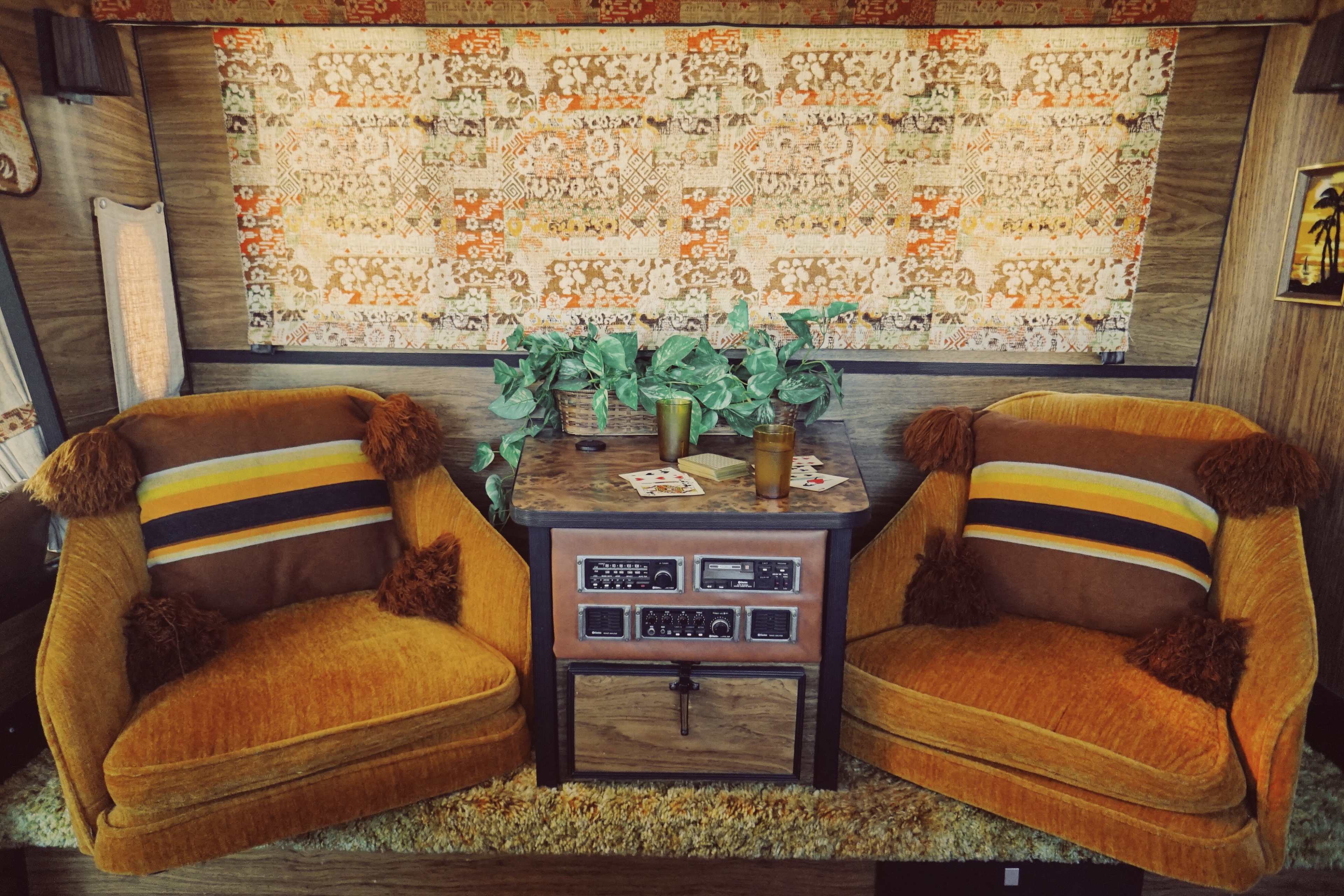 A cozy interior featuring two vintage orange chairs with striped cushions, positioned around a small wooden table adorned with plants and playing cards against a patterned wallpaper backdrop.