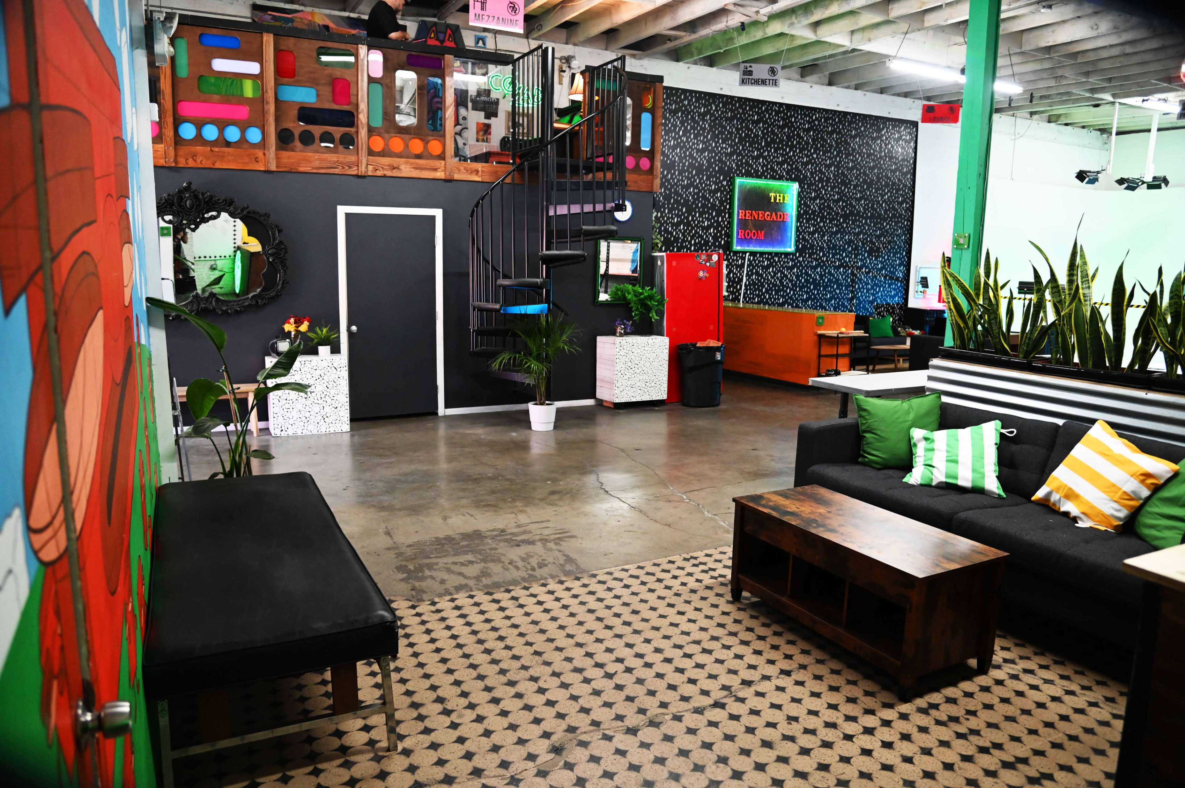 The image shows a modern, spacious indoor area featuring a staircase, seating arrangements, and colorful wall decorations.