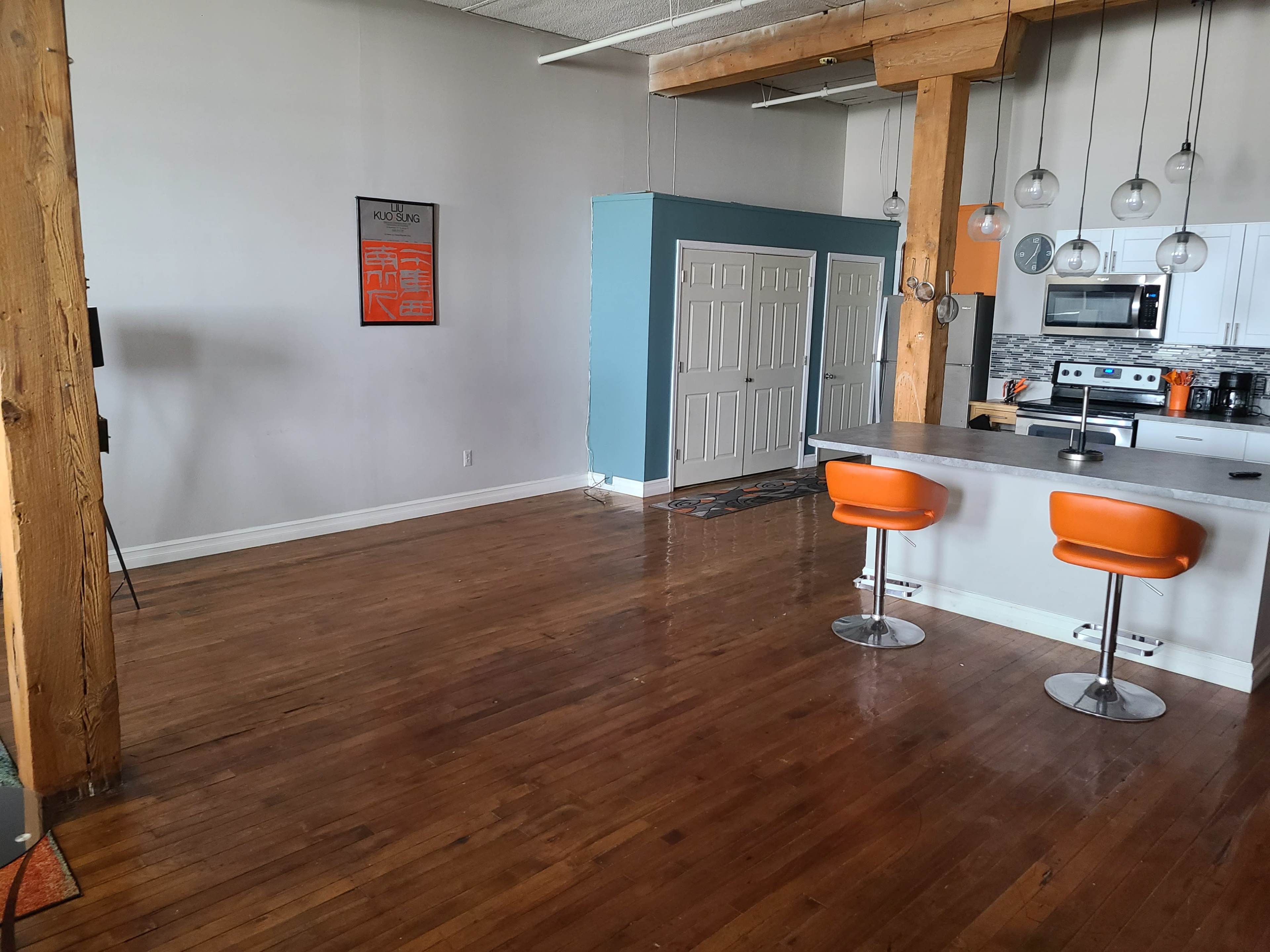 A spacious, empty loft-style living area with hardwood floors, a kitchen area featuring stainless steel appliances, and two orange barstools at a kitchen island.