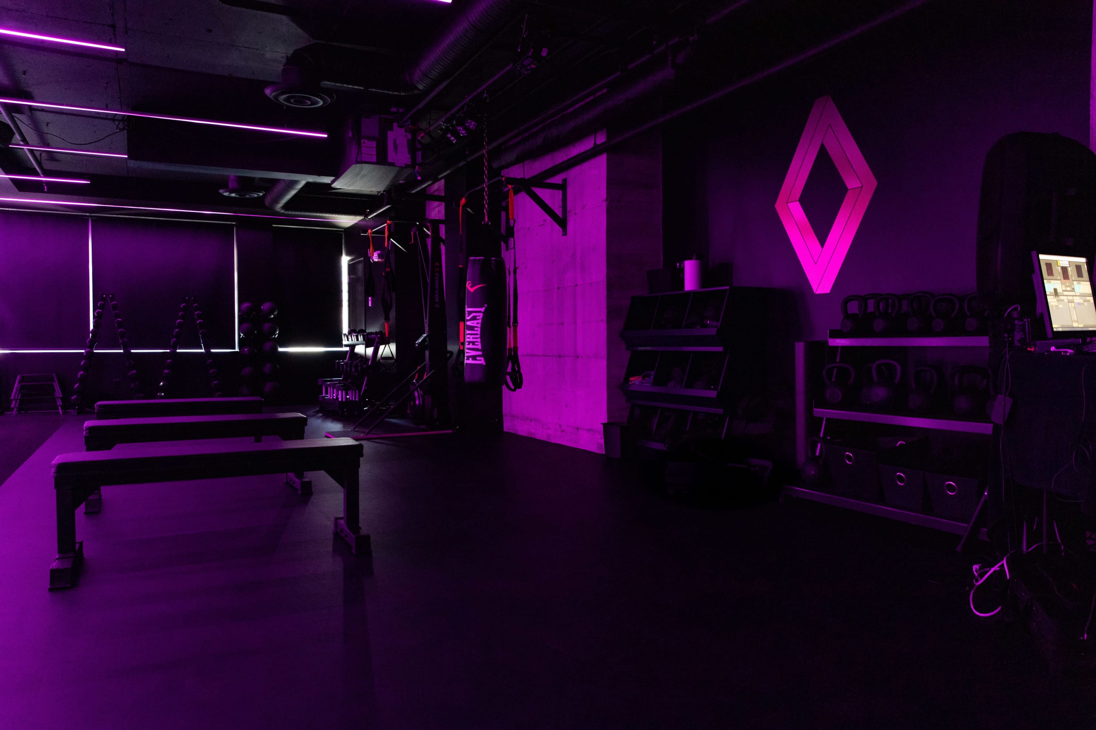 A dimly lit gym with purple lighting, featuring exercise benches and various weights arranged against the walls.