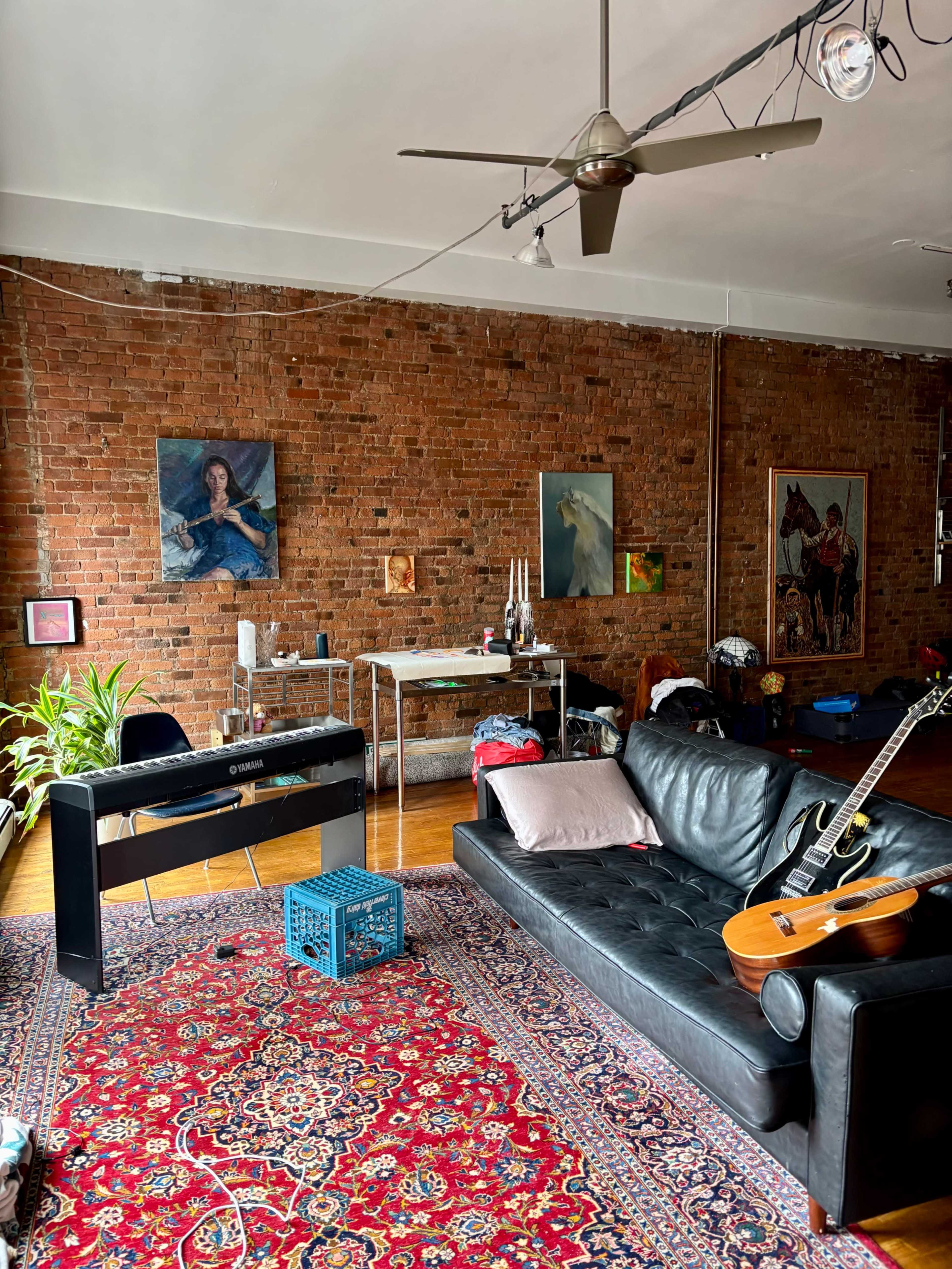 The image shows a living room with exposed brick walls, a black leather sofa, a piano, and a guitar, along with various artworks and plants.