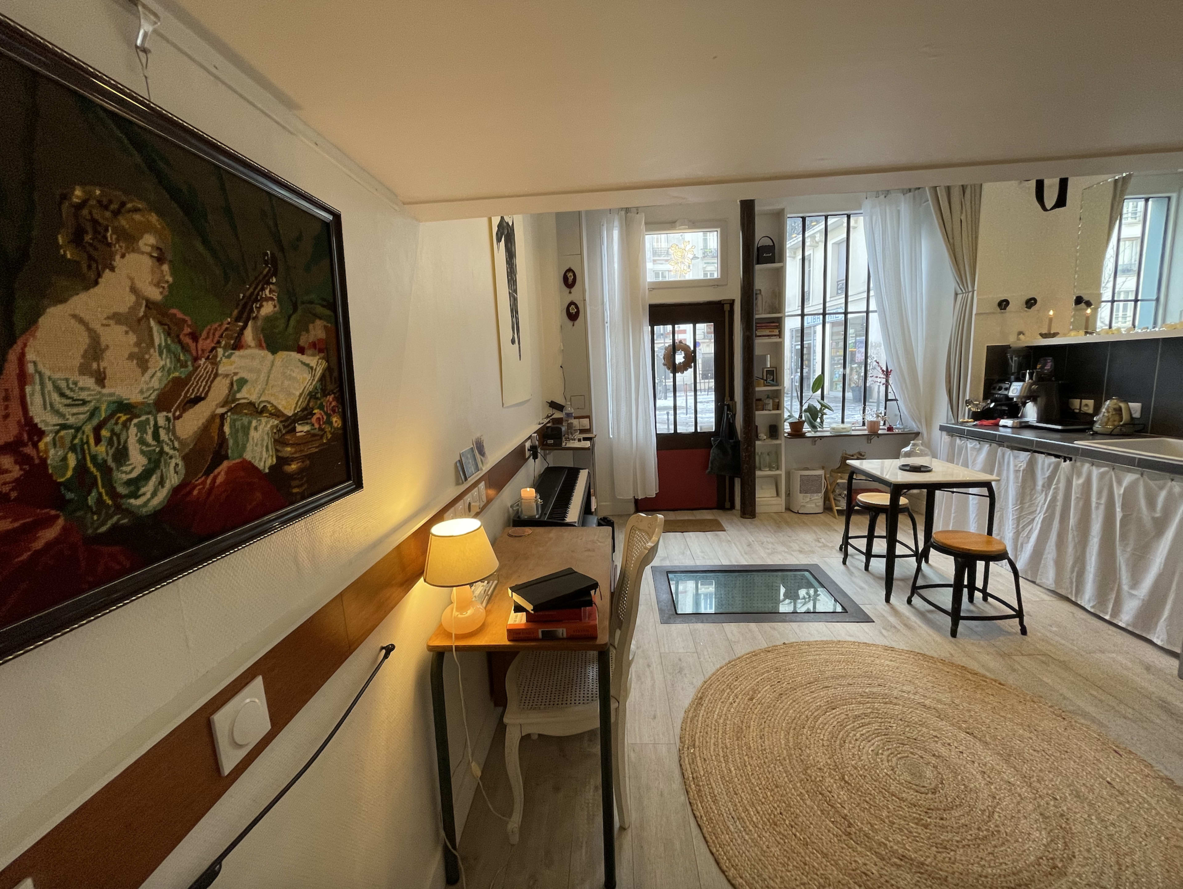 The image shows a cozy interior space with a painting on the wall, a desk with a lamp, a rug on the wooden floor, and kitchen elements in the background.