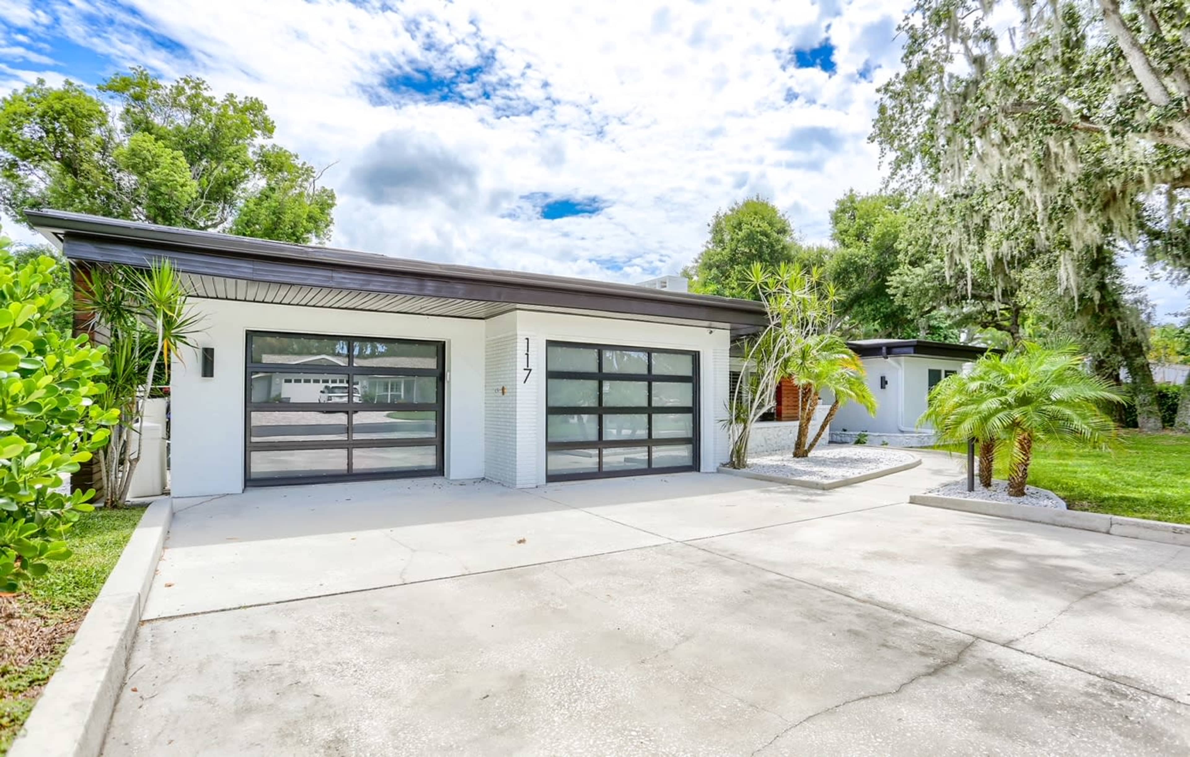 A modern single-story house with a large driveway features a two-car garage and tropical landscaping.
