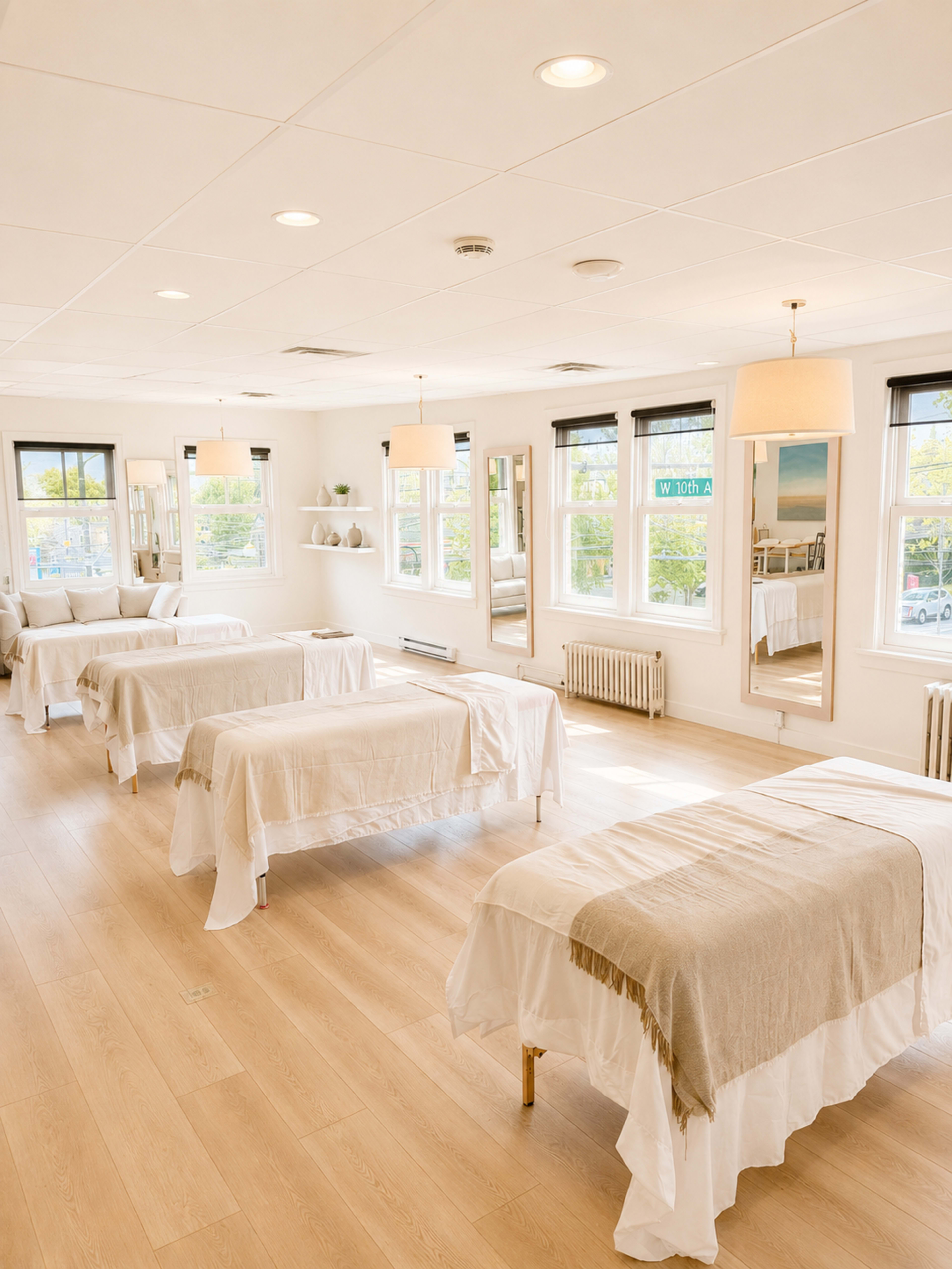The image shows a bright and airy wellness studio with several massage tables arranged neatly, illuminated by natural light from large windows.