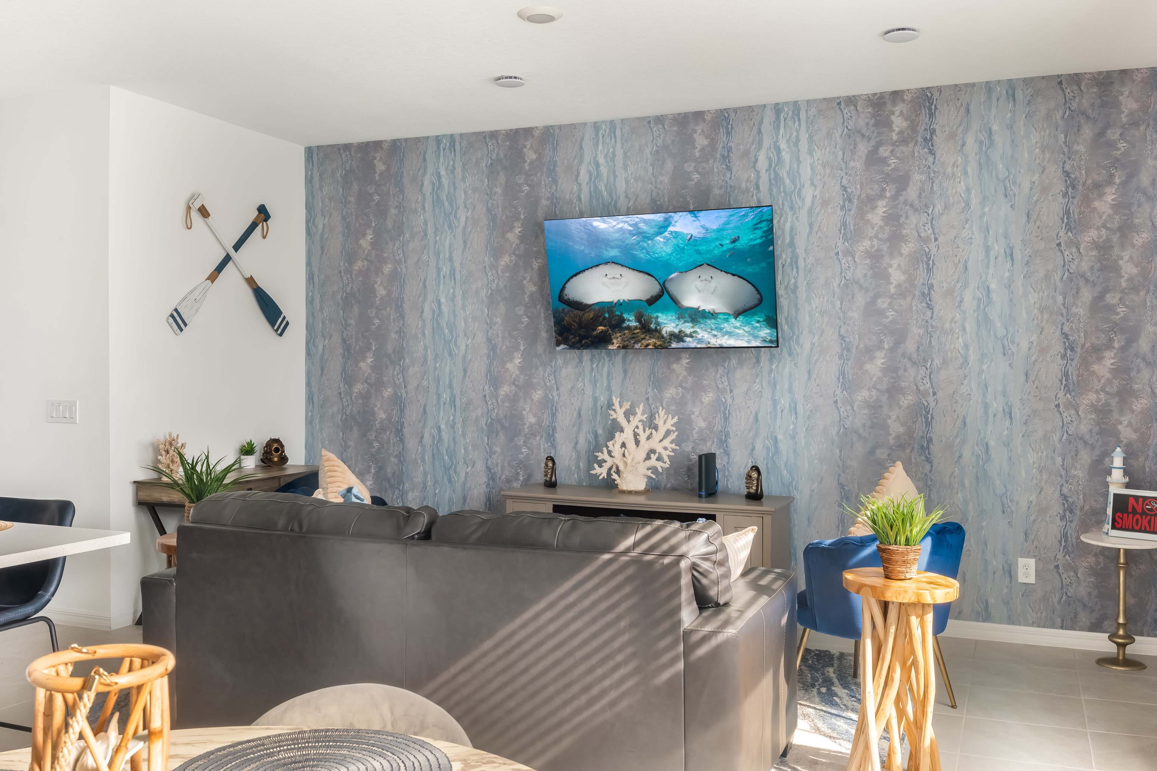 The image shows a modern living room with a dark leather sofa, a wall-mounted television displaying an underwater scene, and decorative elements like plants and marine-themed decor.