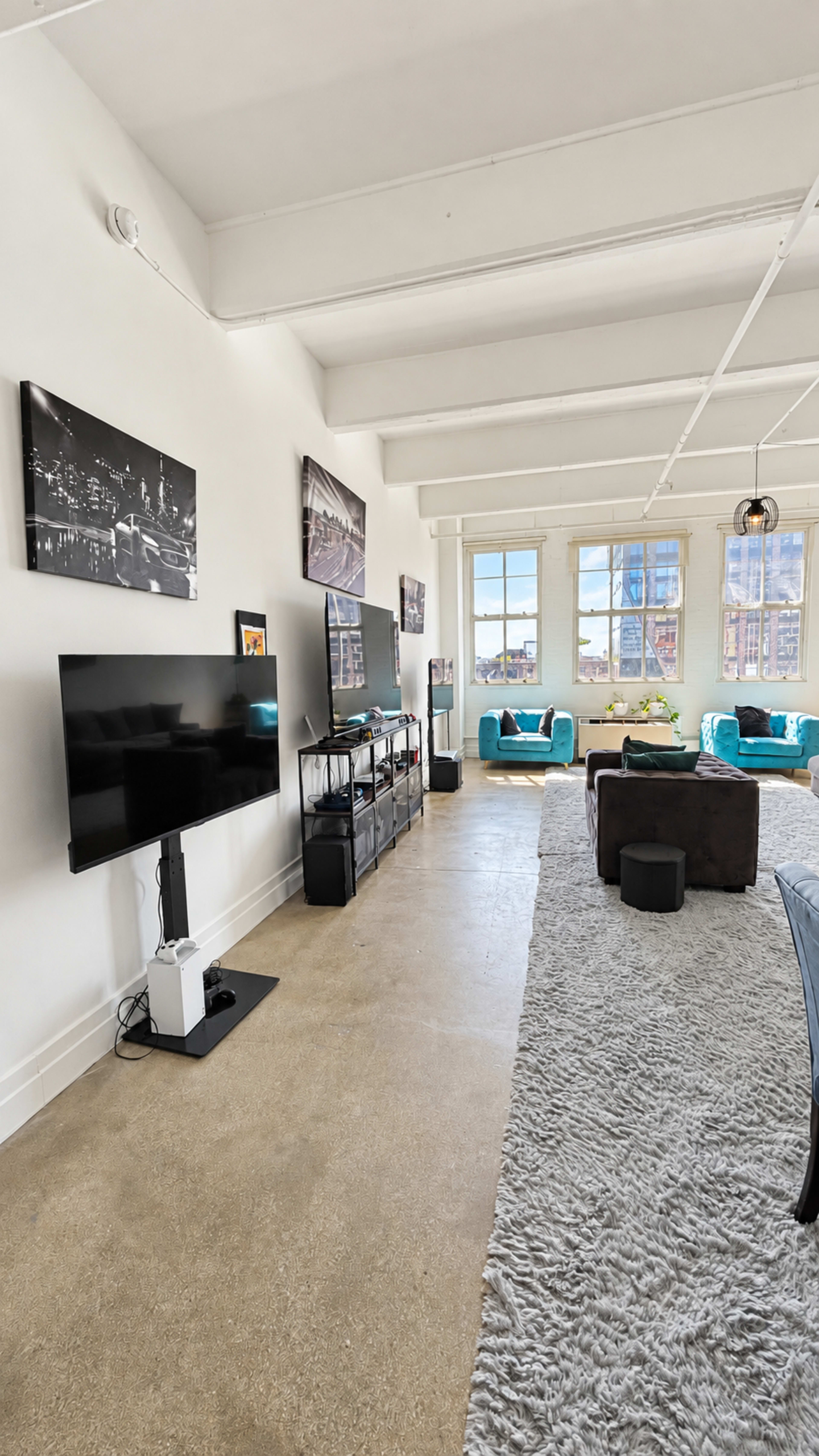 The image shows a spacious, modern living room with a large TV on a stand, several couches in teal and gray, and large windows letting in natural light.