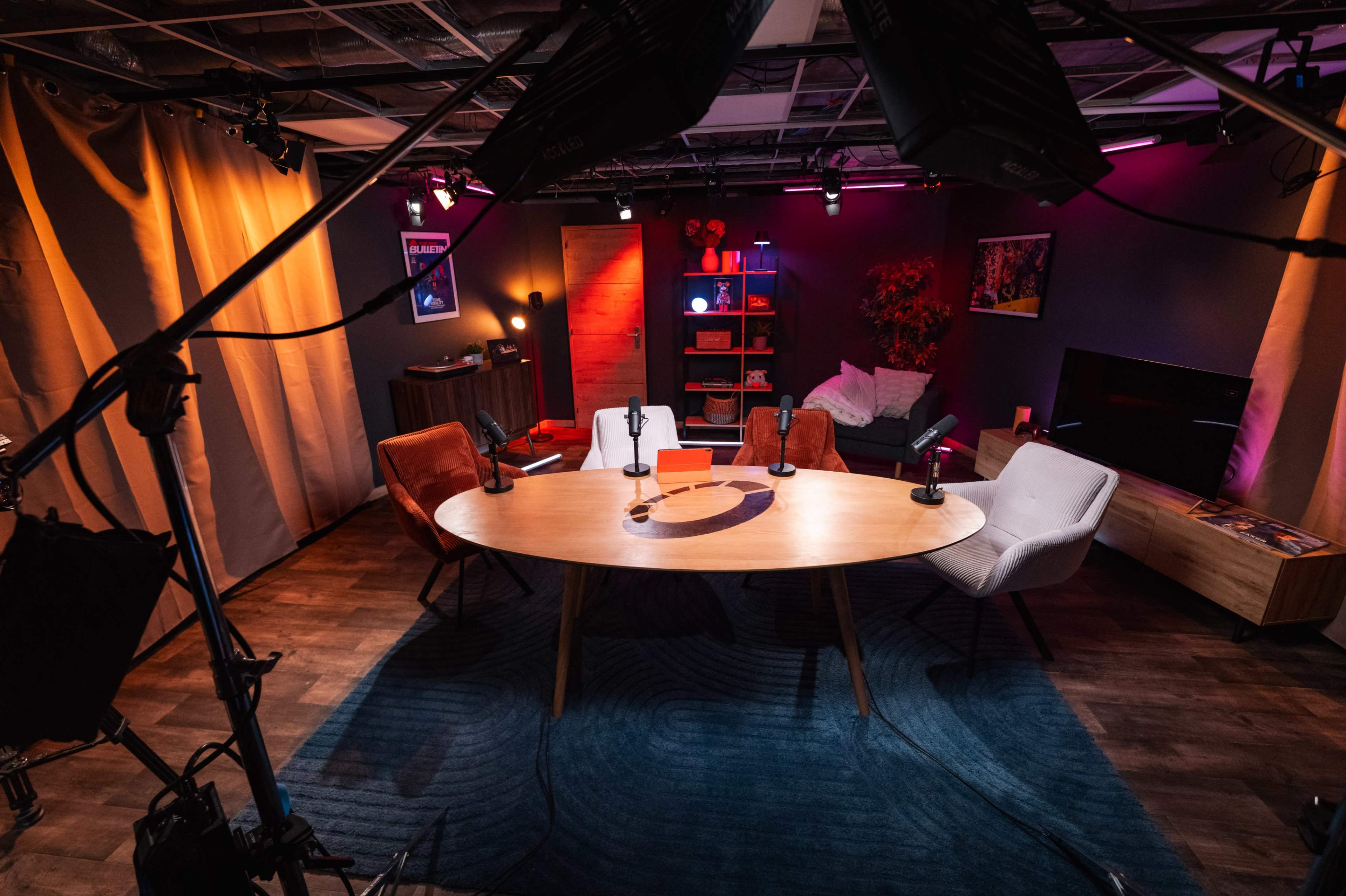 The image shows a modern podcast studio setup with a circular table surrounded by four chairs, microphones positioned on the table, and various decorative elements in the background.