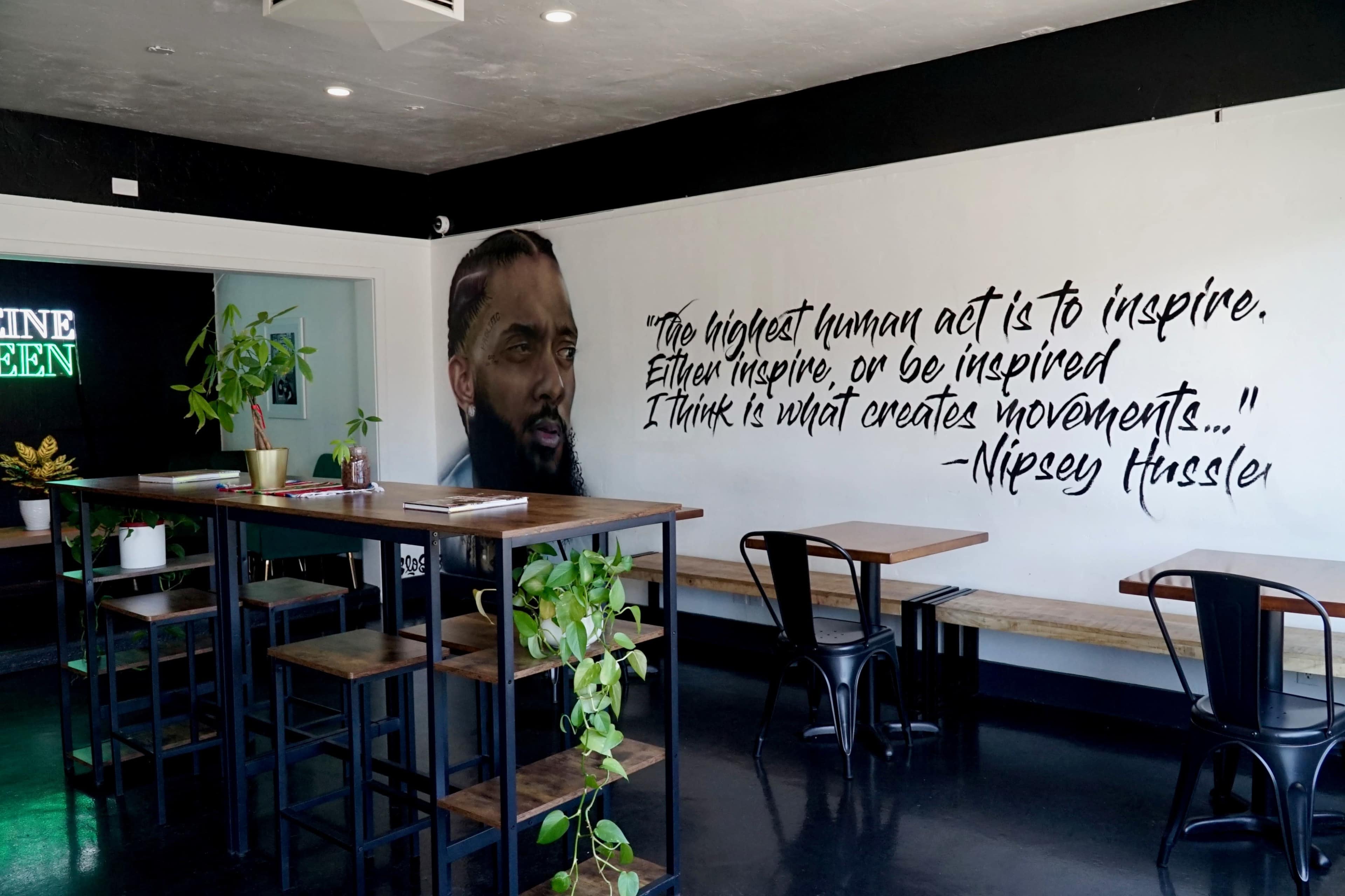 The interior of a café features wooden tables, black chairs, and a large mural of a man with an inspirational quote by Nipsey Hussle on the wall.