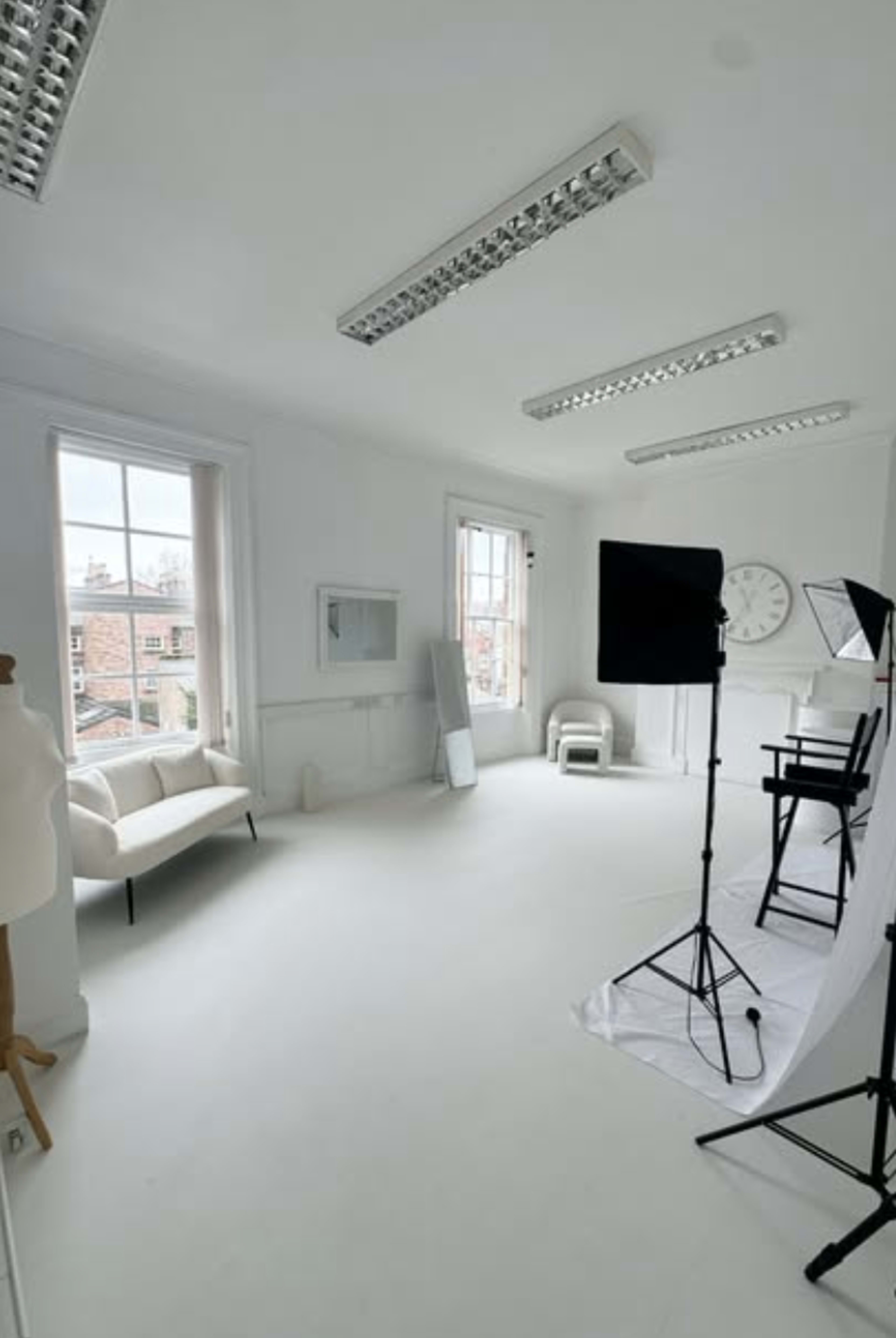The image shows a spacious, well-lit studio with white walls, a white floor, and various photography equipment set up, including light stands and a reflector.