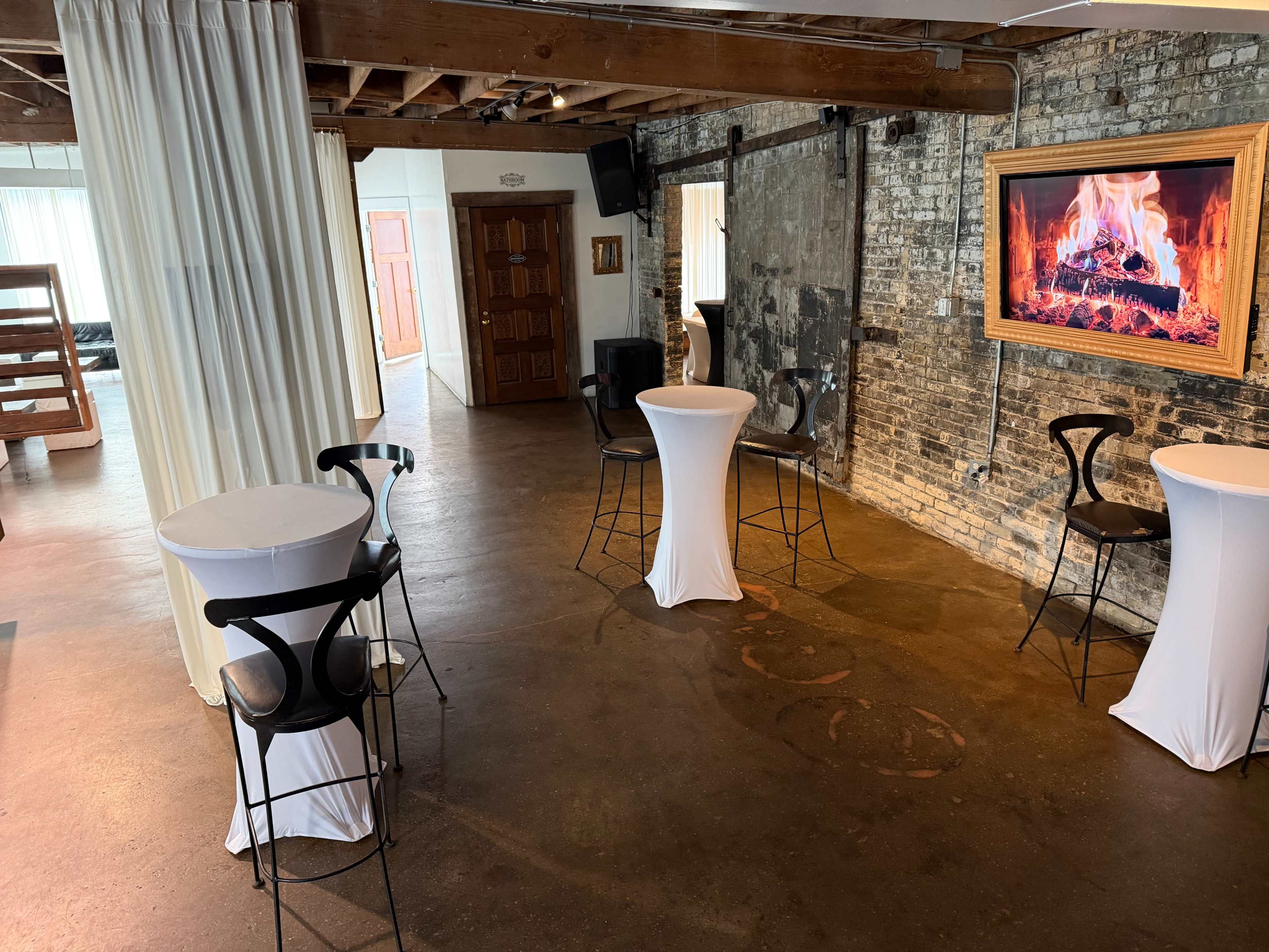 The interior features a spacious venue with high ceilings, exposed brick walls, and several white cocktail tables arranged around a decorative fireplace.