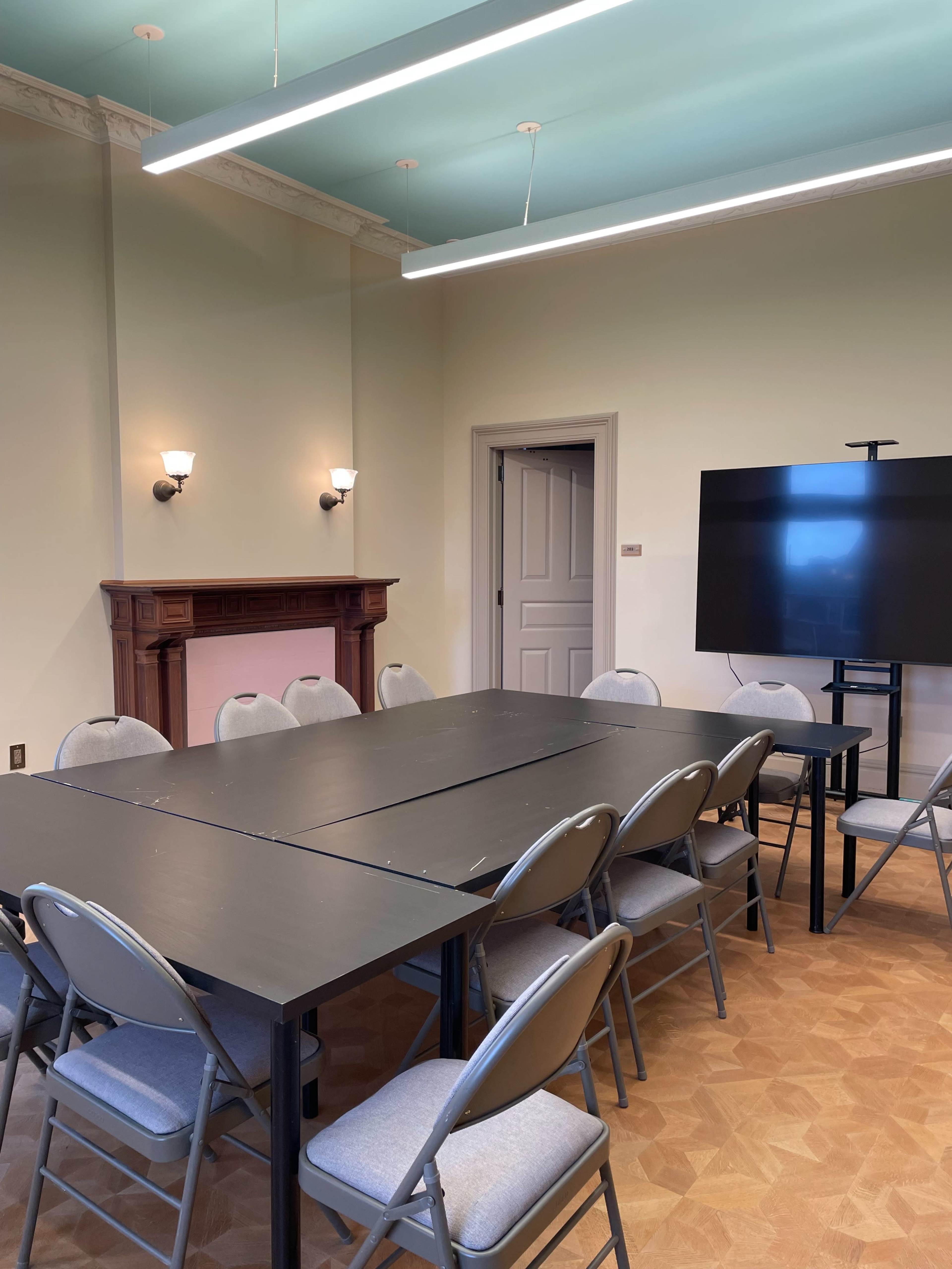 A rectangular black conference table with chairs surrounds it in a well-lit meeting room, featuring a decorative fireplace and a large screen on one side.