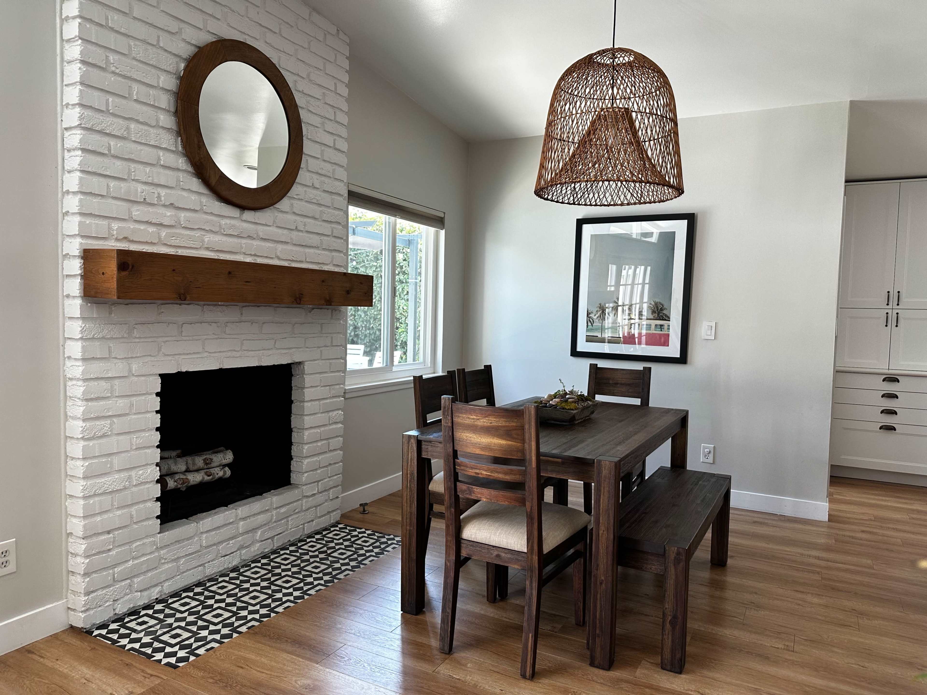 A modern dining area features a wooden table and benches, a white brick fireplace, and a woven pendant light hanging above.
